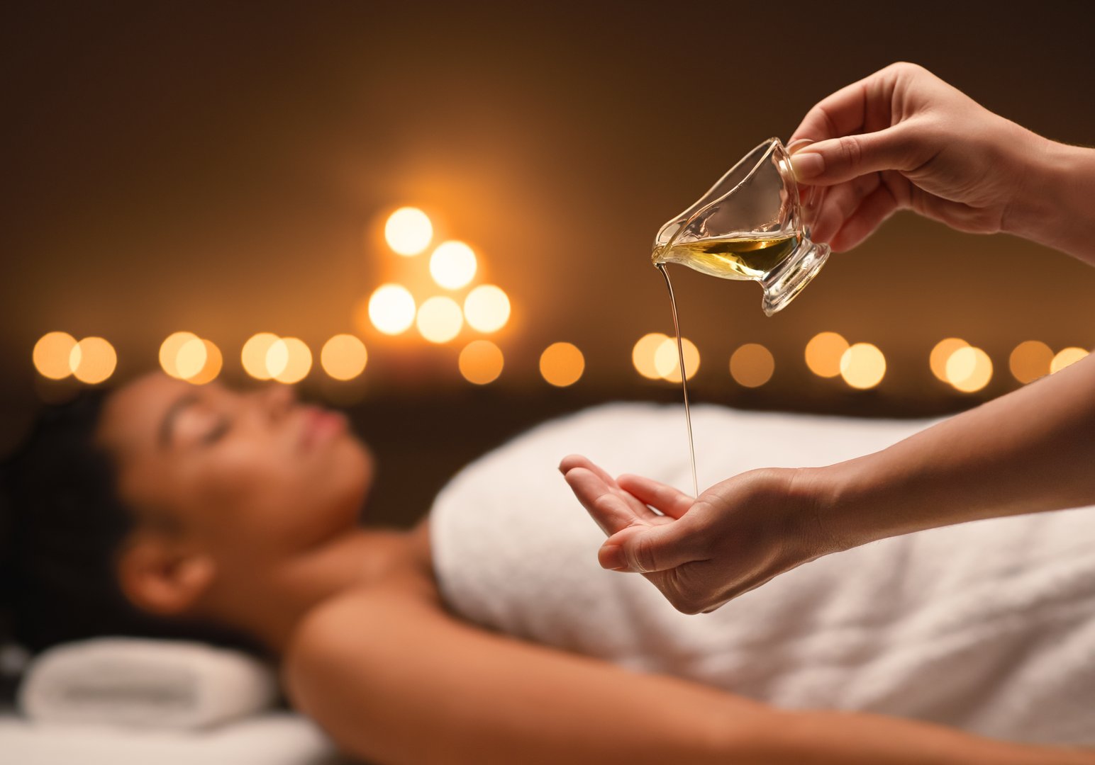 Close up of female therapist applying massage oil on hands before therapy over black lady lying on massage table