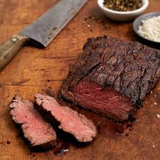 Sliced grilled steak on a wooden cutting board with a large knife and small bowls of salt and pepper in the background.