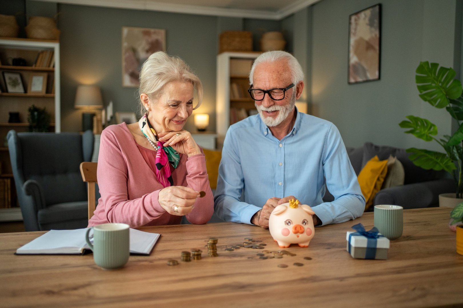 Smiling senior couple sitting at a wooden table, joyfully placing coins into a piggy bank, celebrating their efforts in managing finances and planning for a secure retirement