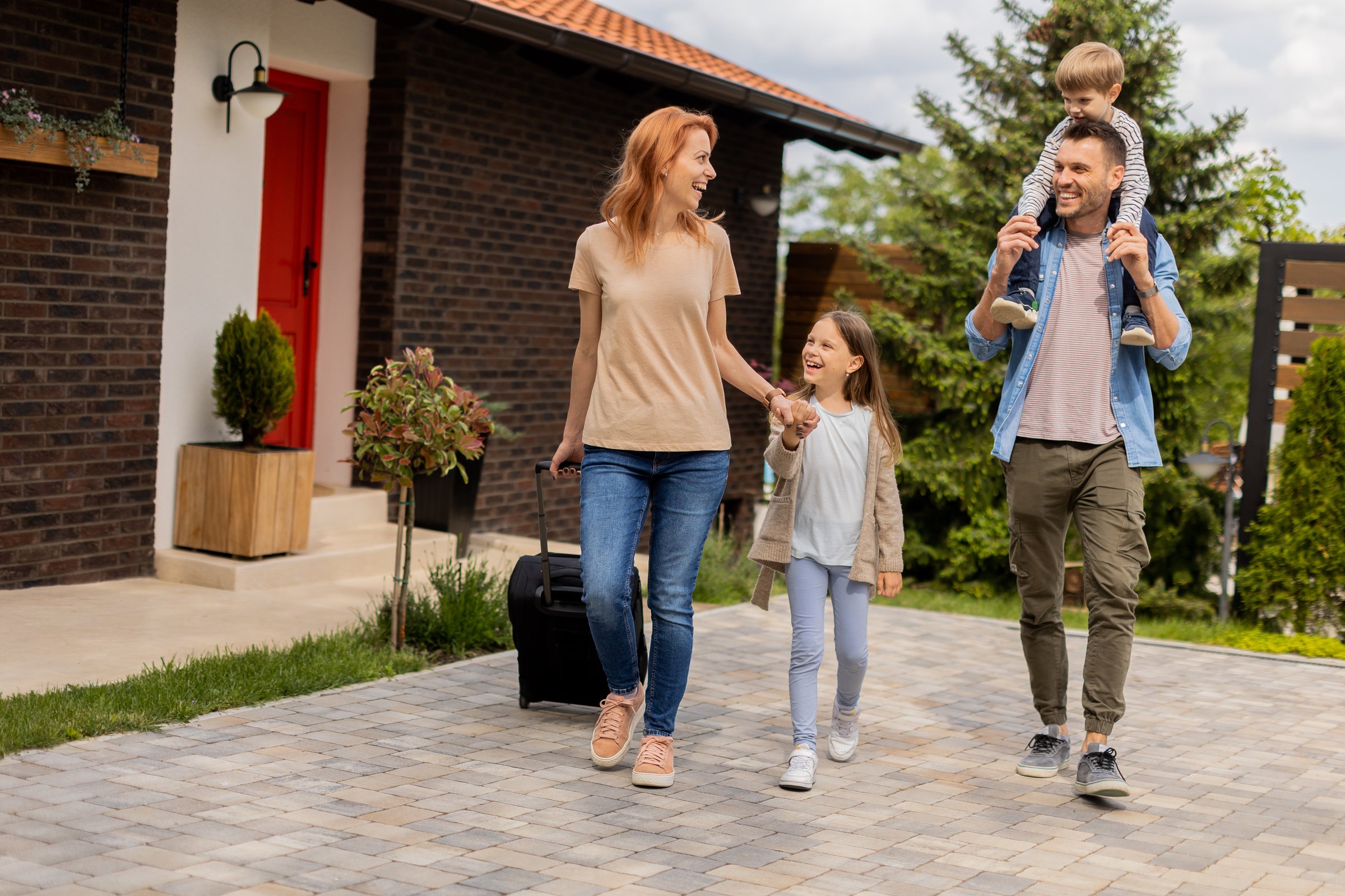 Family with a mother, father, son and daughter walking with  abaggage outside on the front porch of a brick house