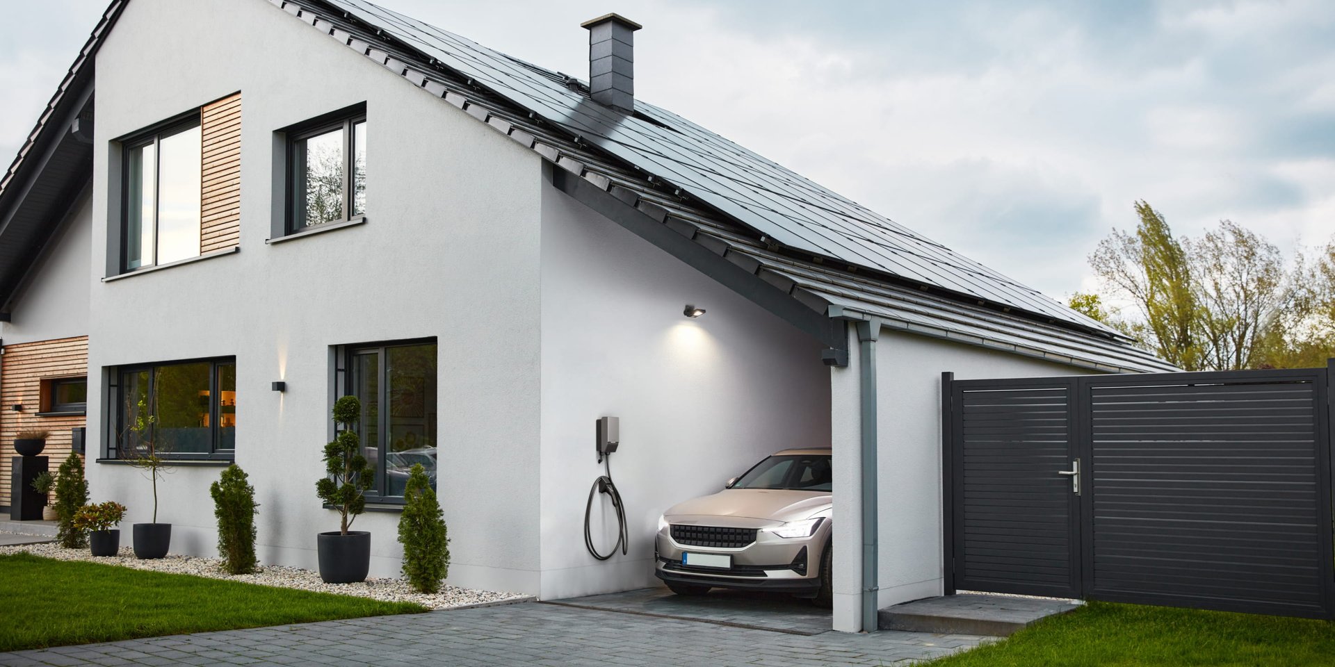 Modern house with solar panels, a car parked in the driveway, and a charging station. Neatly landscaped yard.