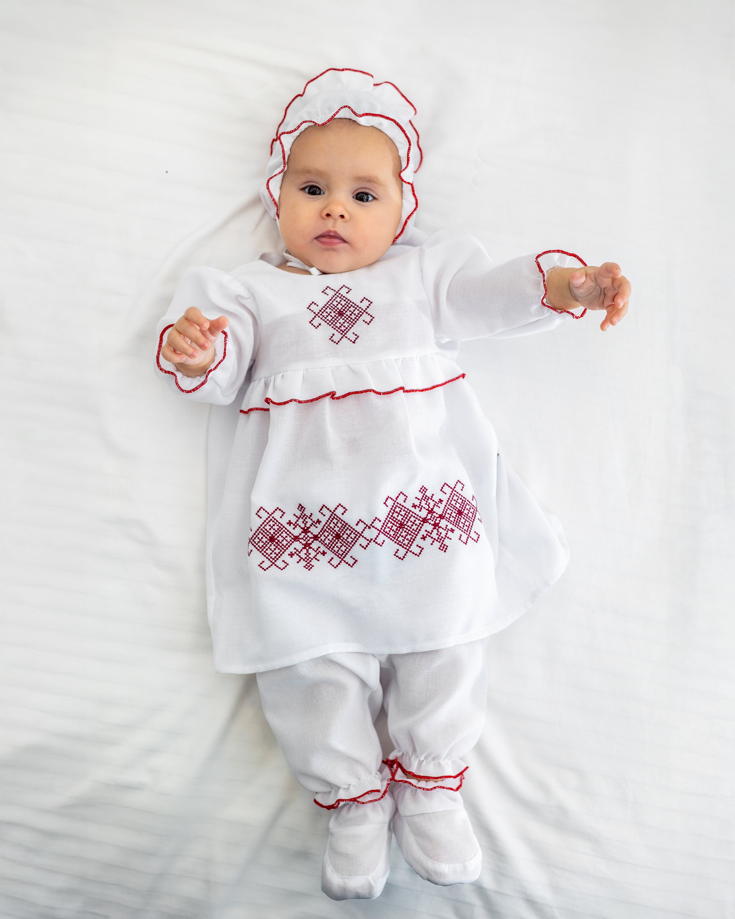 A baby girl dressed in a white embroidered dress lies on a soft white bed. Traditional infant clothing highlights purity, innocence and tenderness of early childhood. Image represents baby fashion, christening outfit, family values, newborn lifestyle and calm minimalist aesthetic.