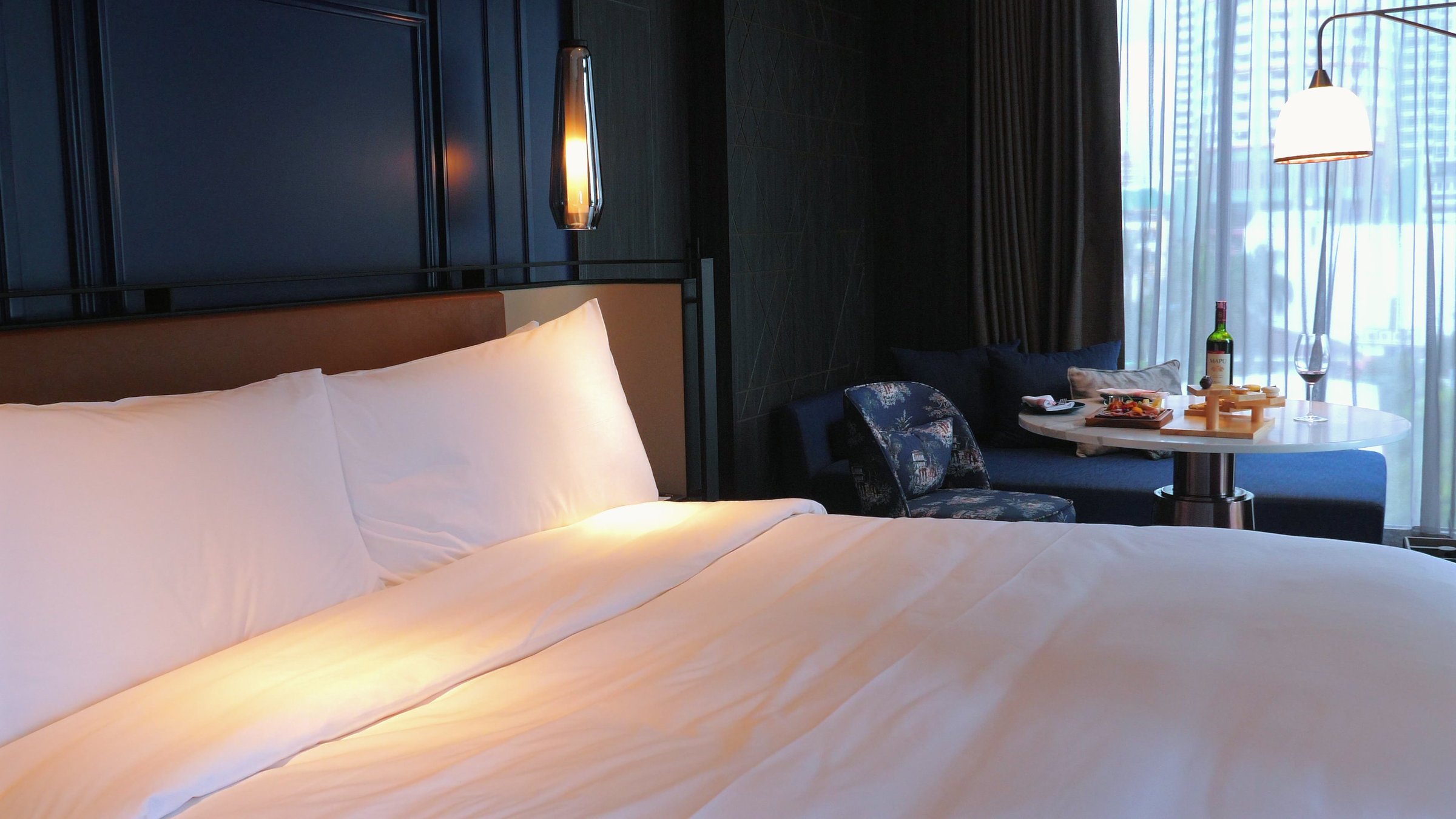 Cozy modern bedroom in luxury hotel with comfortable king-size bed, bottle of wine and snacks on table by window, perfect for relaxation and unwinding. Tranquility and Comfort.