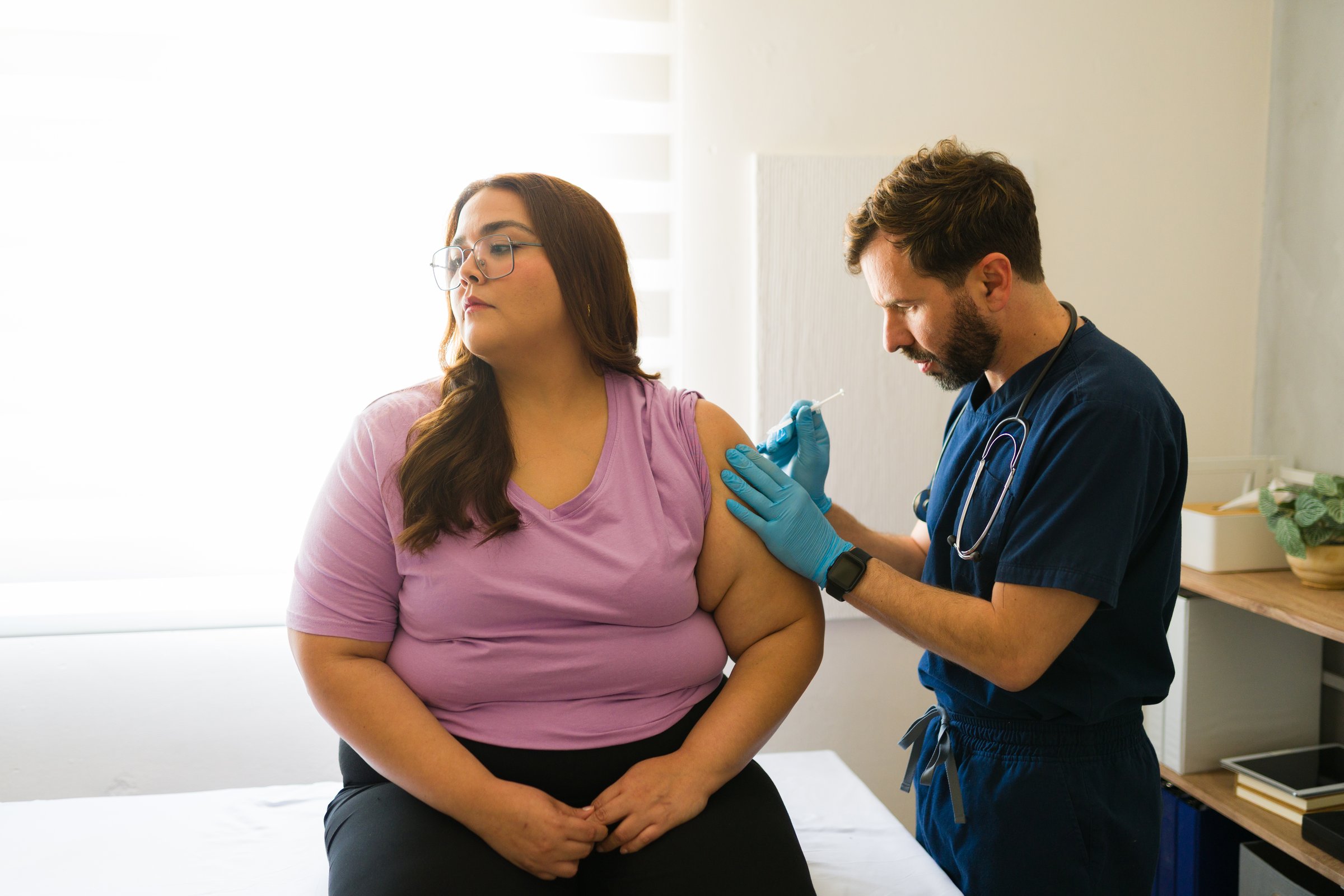 Caucasian male doctor injecting a vaccine into the arm of a plus-size female patient during a medical appointment at the clinic