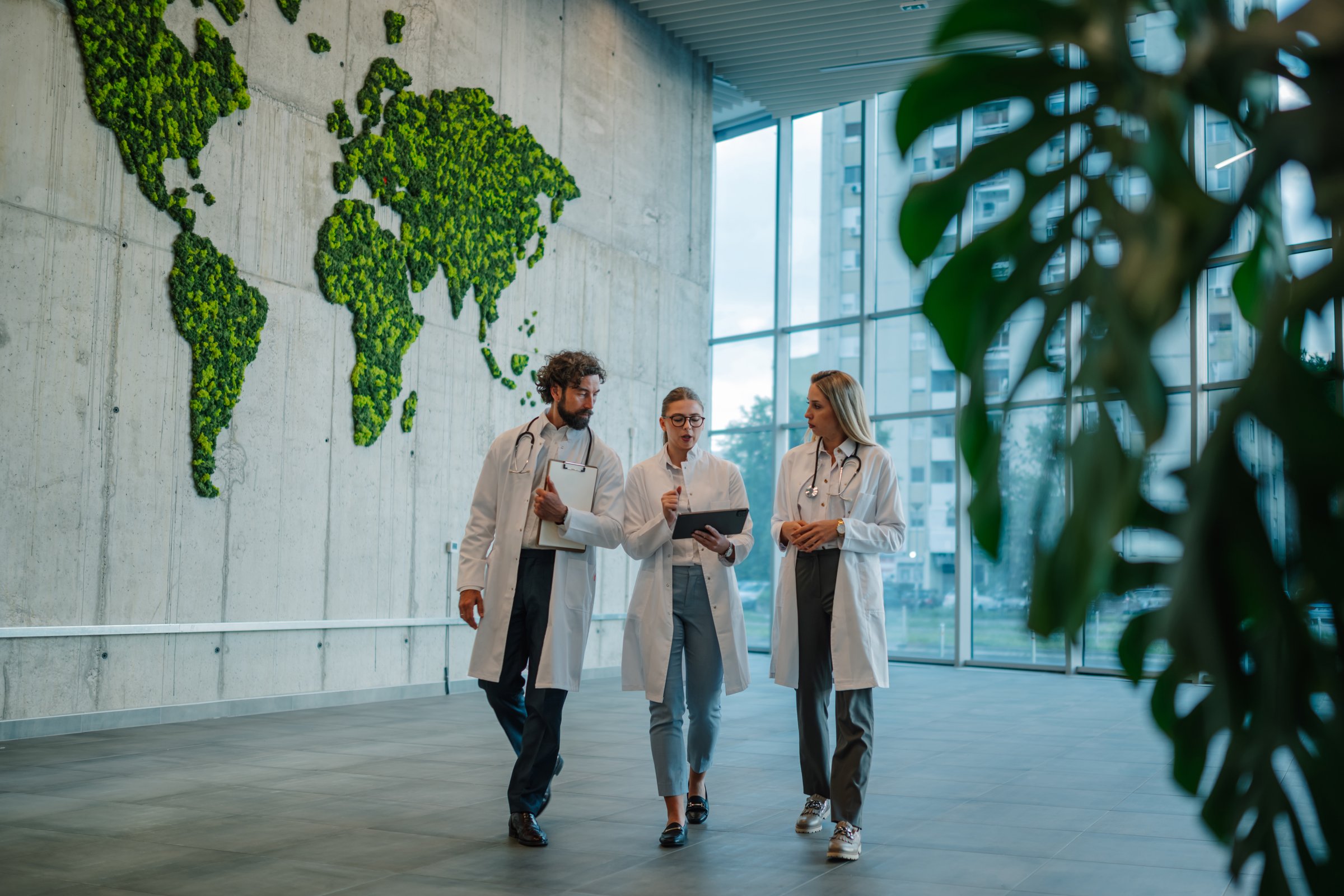 Three doctors are walking and discussing a diagnosis in a modern hospital lobby with a vertical garden shaped like a world map