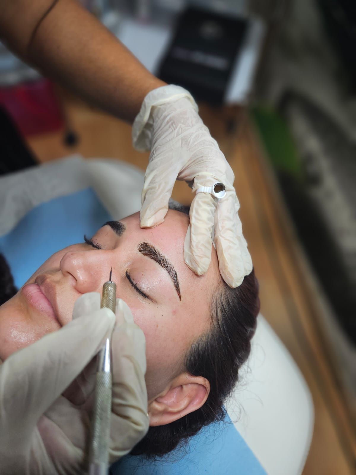 Person receiving eyebrow microblading treatment, with gloved hands holding a tool near their eyebrow.