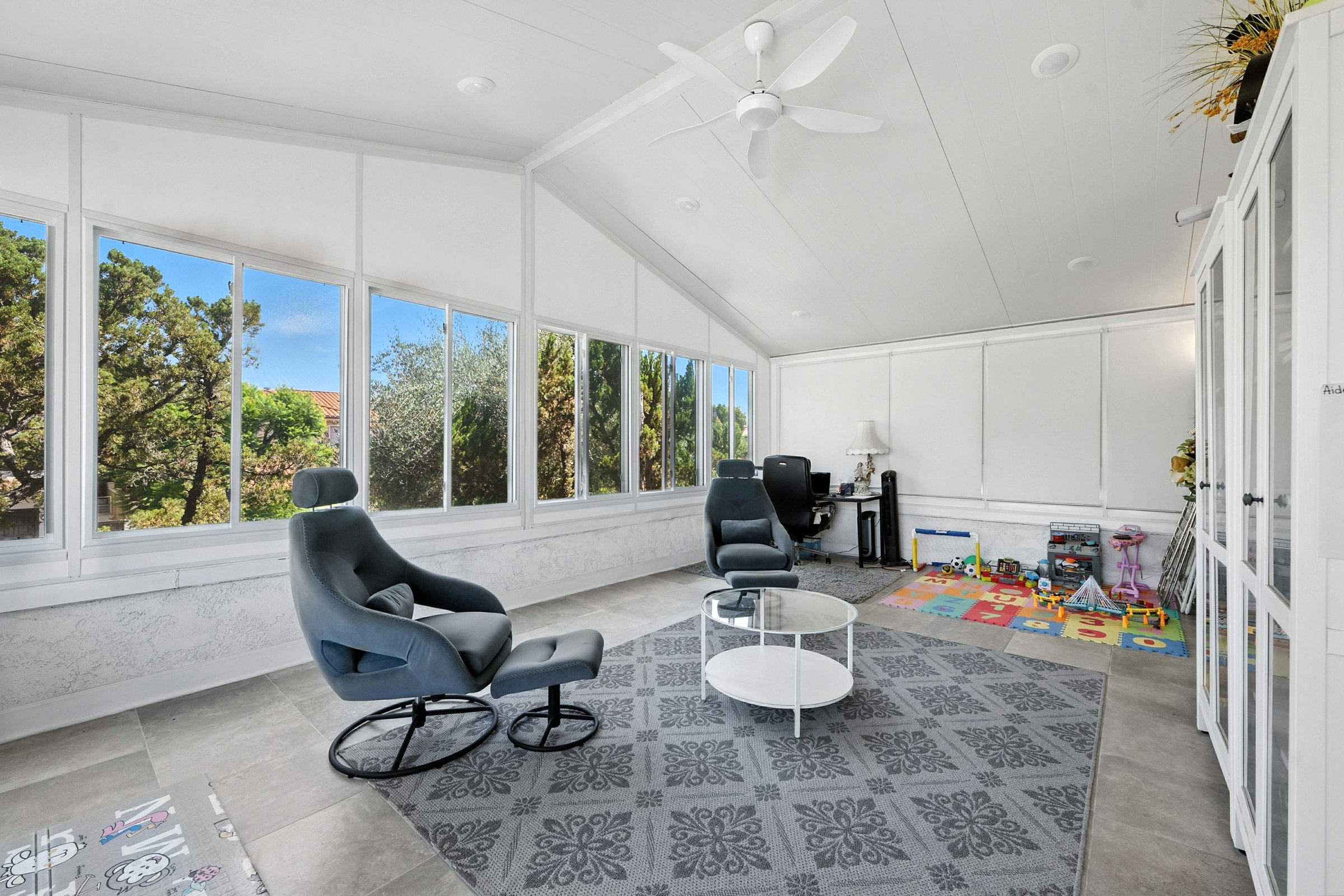 A sun-drenched sunroom features comfortable gray armchairs, a geometric rug, and ample windows offering views of lush greenery.