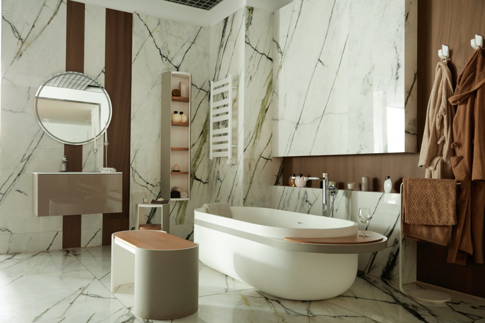 Modern bathroom featuring freestanding bathtub, marble walls, round mirror, wooden accents, shelving with decorative objects, towel rack, bathrobes hanging on wall hooks