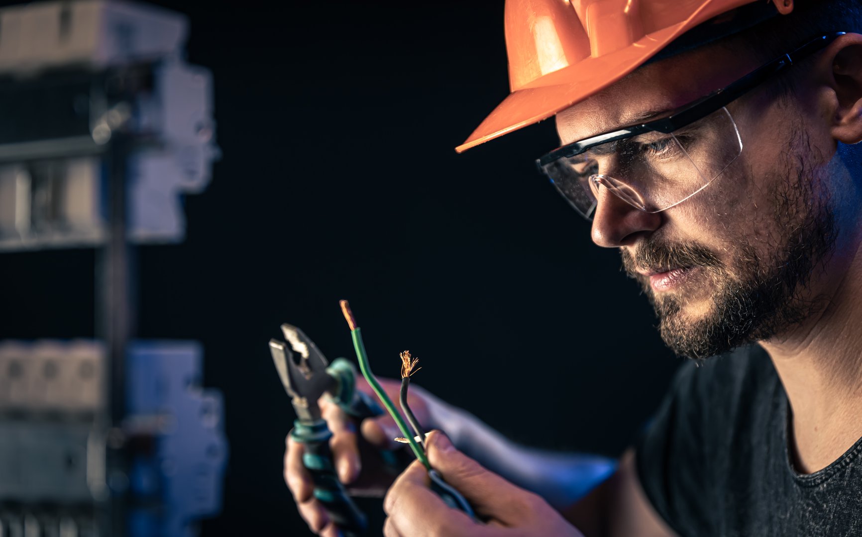A male electrician in a protective helmet works in a switchboard with an electrical connecting cable.