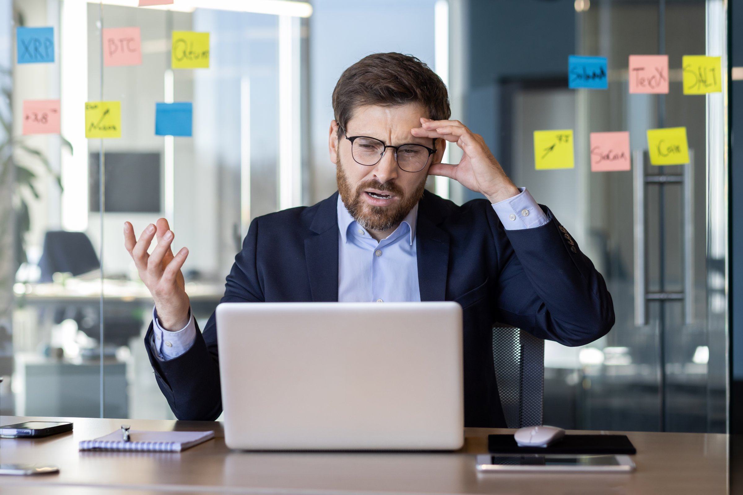 Businessman feeling frustrated while working at office desk with laptop. Notes on glass wall in background. Concept of stress, frustration, and business challenges