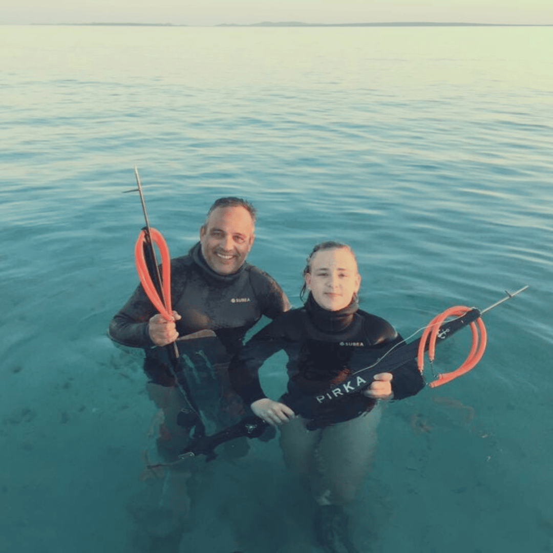 Two people in wetsuits holding spearguns in clear, shallow water, smiling at the camera.