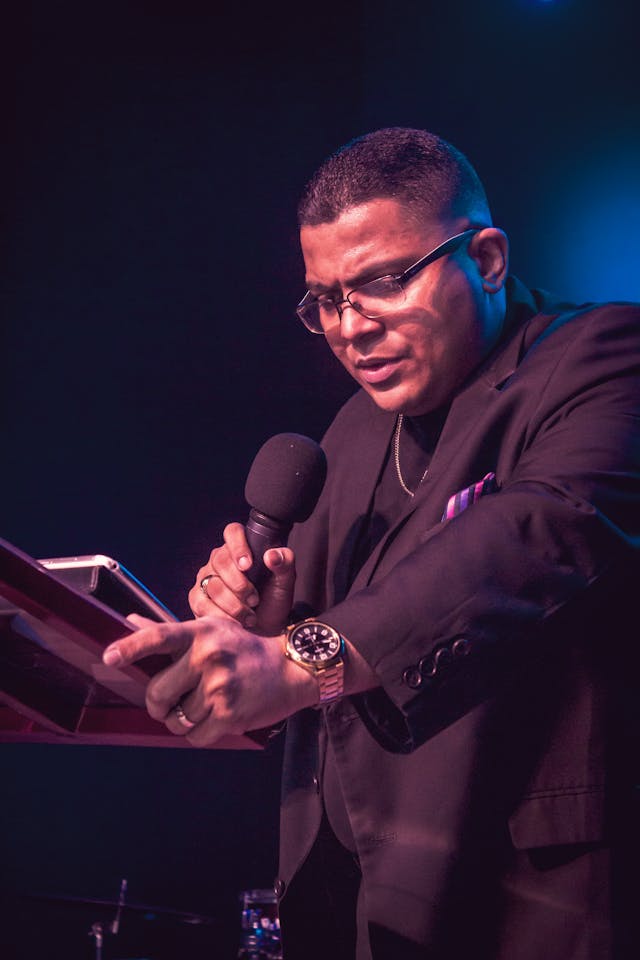 Man in suit holding a microphone, reading from a book on a stand, speaking passionately under stage lighting.
