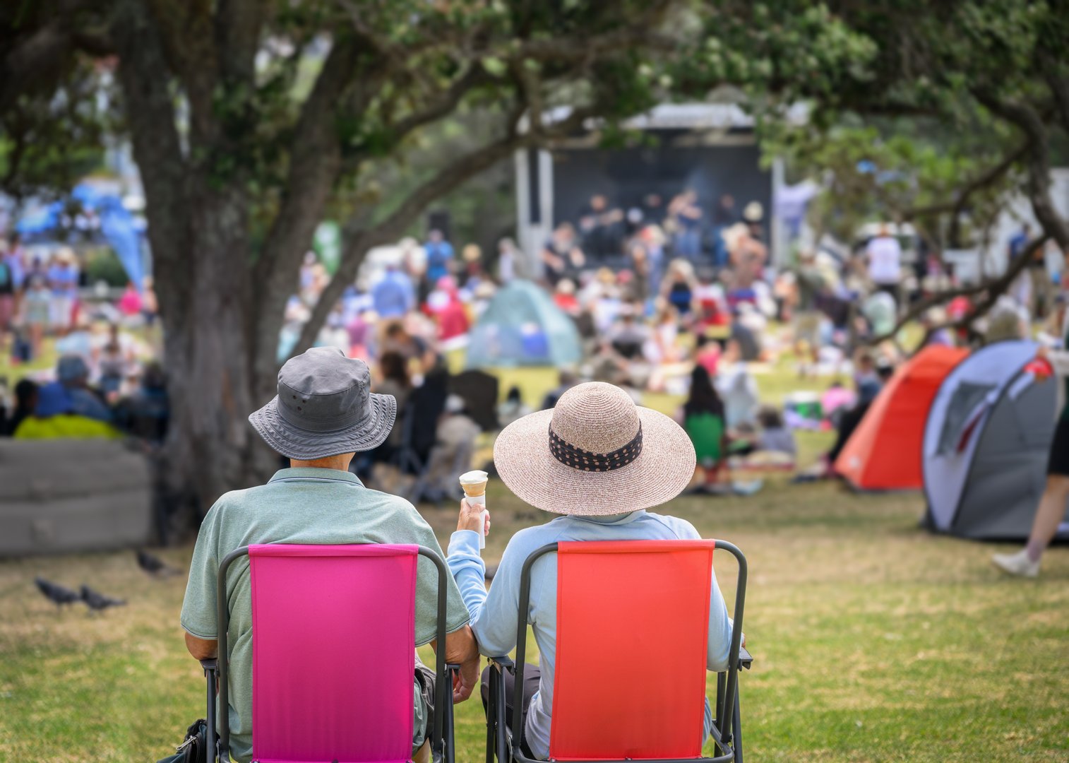 Couple sitting on camping chairs, unrecognizable crowds listen to the music concert in the park. Auckland.