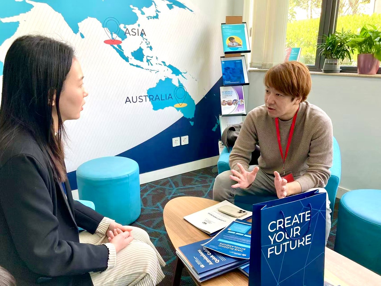 Two people seated in a modern office, discussing near a "Create Your Future" sign with a world map on the wall behind them.