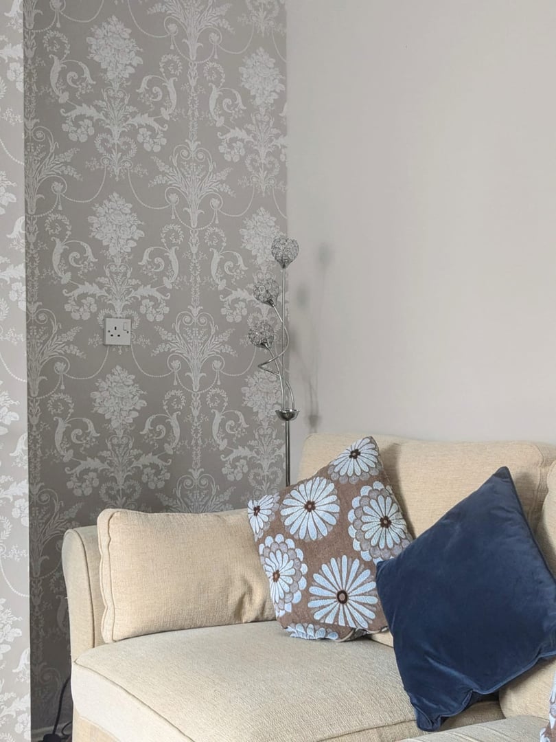 A beige sofa with blue and floral cushions against a wall with ornate floral wallpaper and a floor lamp.
