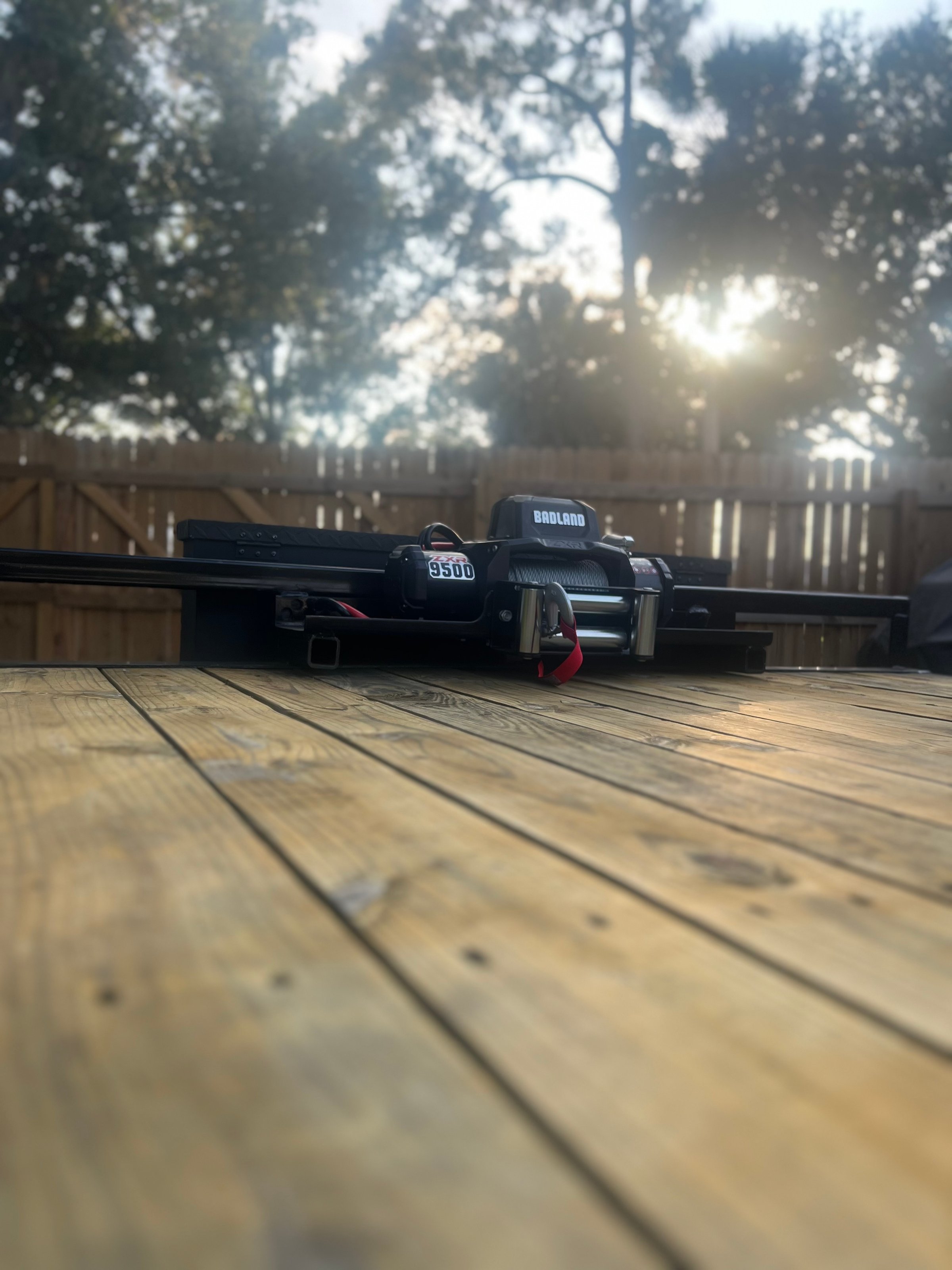 A winch mechanism on a wooden deck outdoors, with trees and sunlight in the background.
