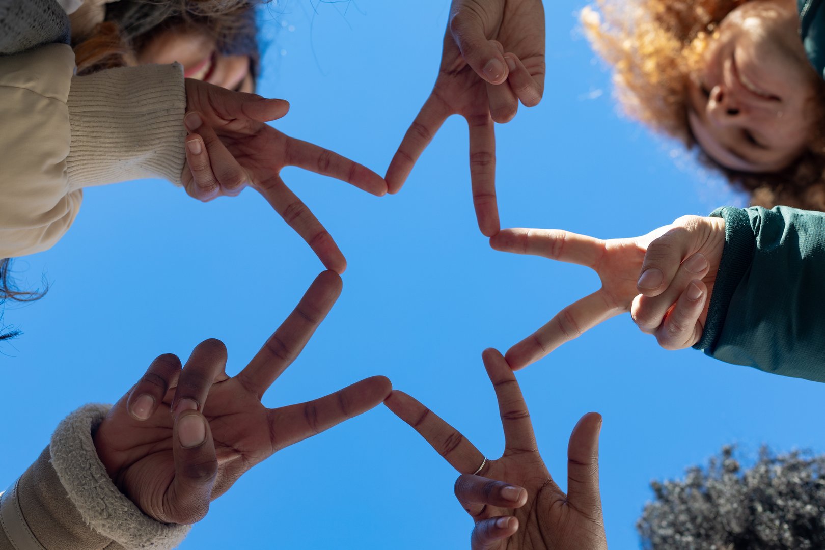 Friends are joining hands, creating a star symbol of unity and teamwork against a blue sky