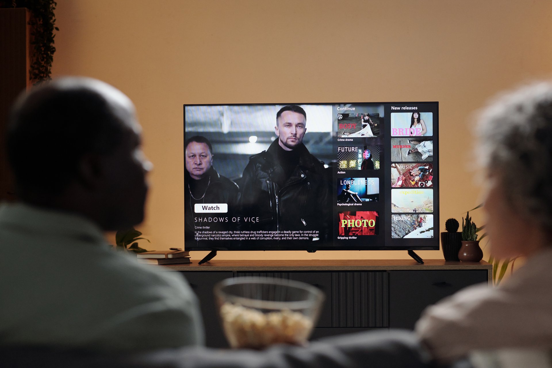 Two people sitting on couch watching action movie on flat-screen TV featuring action-packed scenes and menu screen with various movie options visible