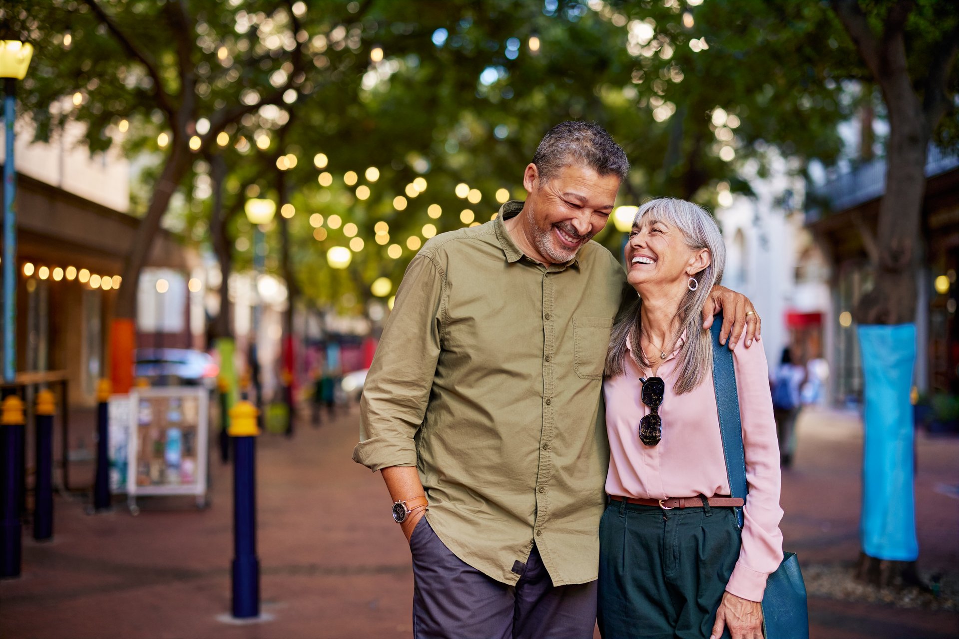 Smiling older couple walks together on a city street. Senior man embracing beautiful woman enjoying a cheerful moment outdoors with copy space. Older couple in love laughs while walking together under string lights in town.
