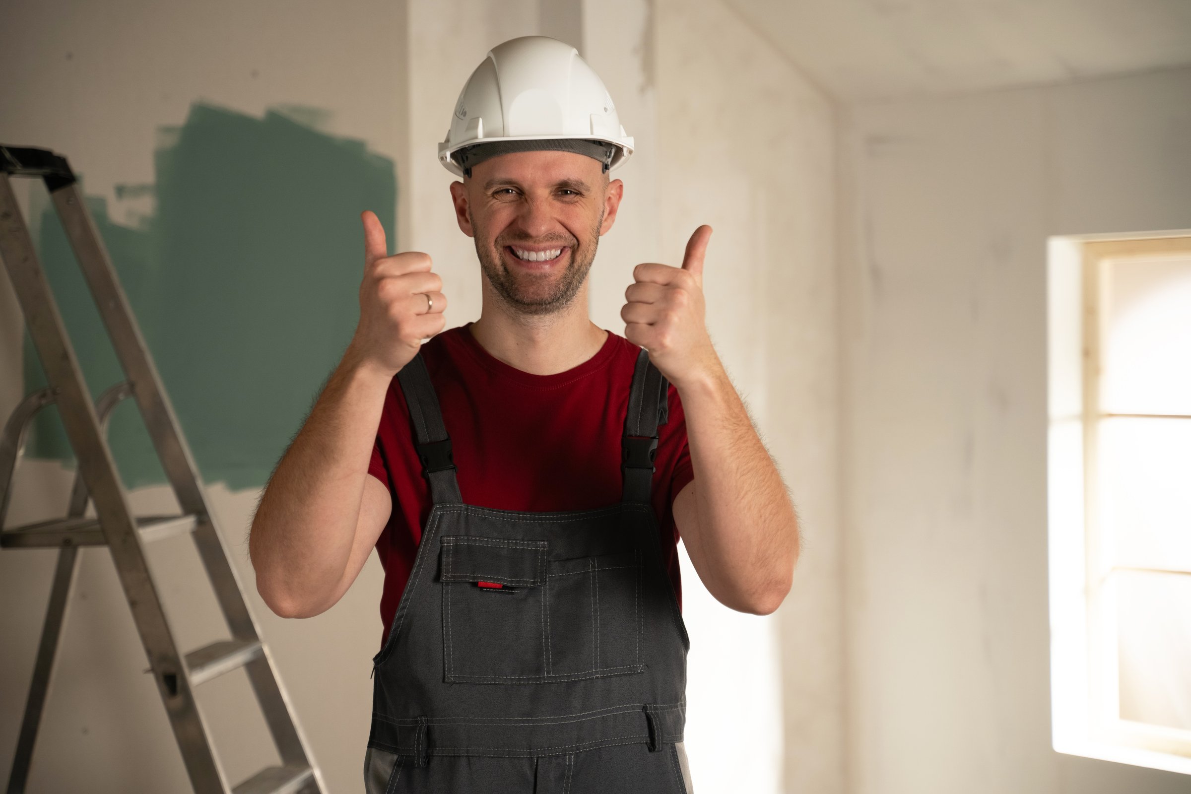 Happy builder wearing work overalls and hardhat showing thumbs up gesture, satisfied with house renovation progress, standing in room with ladder and partially painted wall