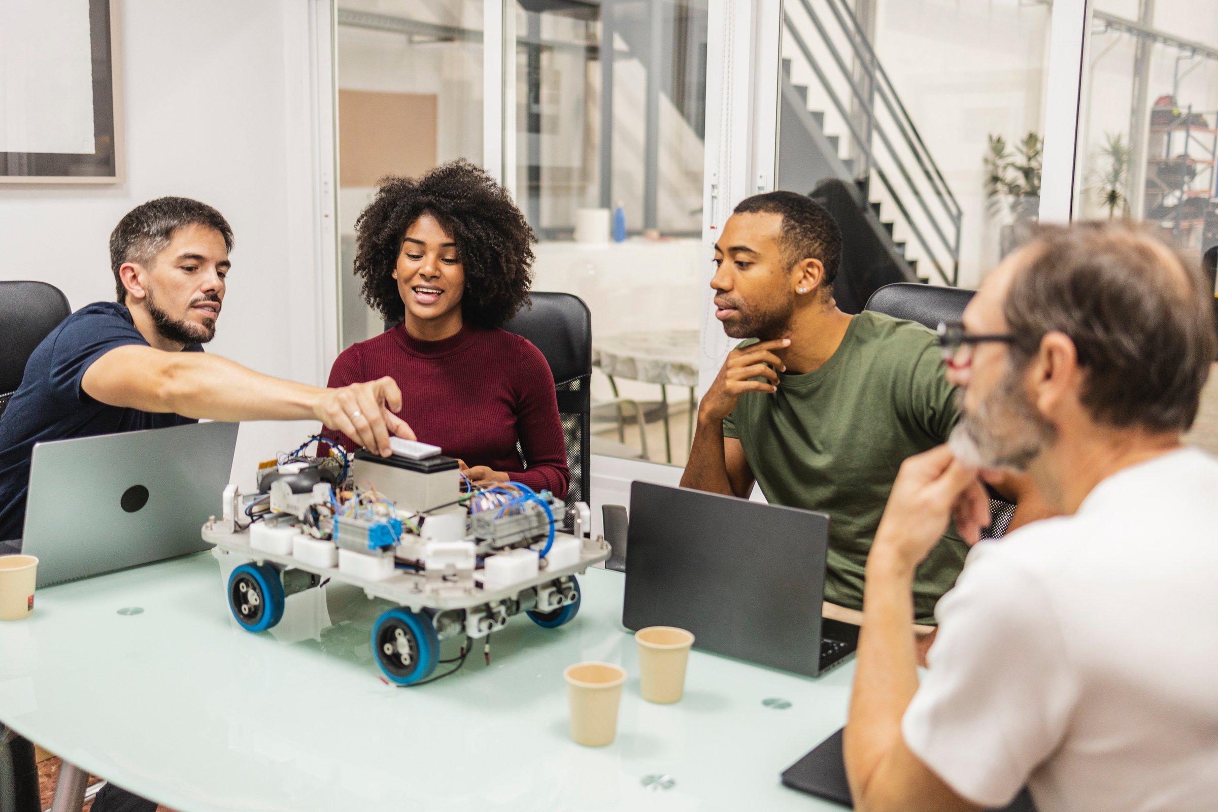 Four engineers, men and women, are discussing a robotics project in their modern office, collaborating and working together around a table with laptops and a prototype robot