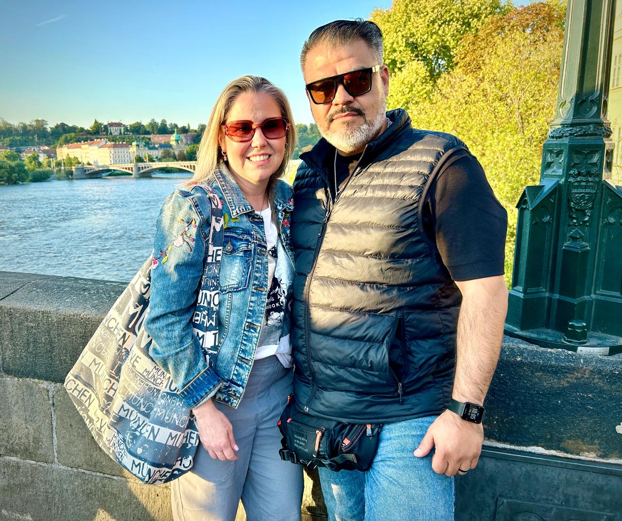 A couple posing together on a riverside path with a bridge and trees in the background, both wearing sunglasses.