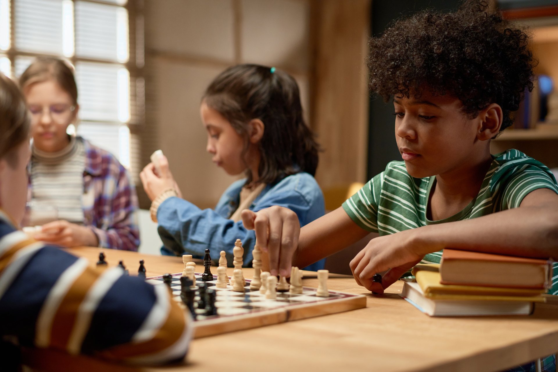 Clever African American schoolboy moving chess piece on board while sitting by table in front of his friend and playing with him