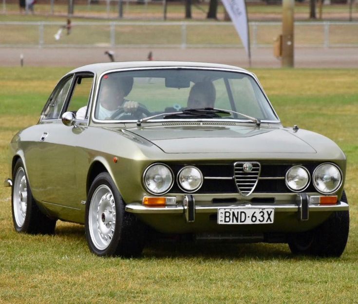 Vintage green Alfa Romeo coupe parked on grass with two people inside, displaying a license plate BND-63X.
