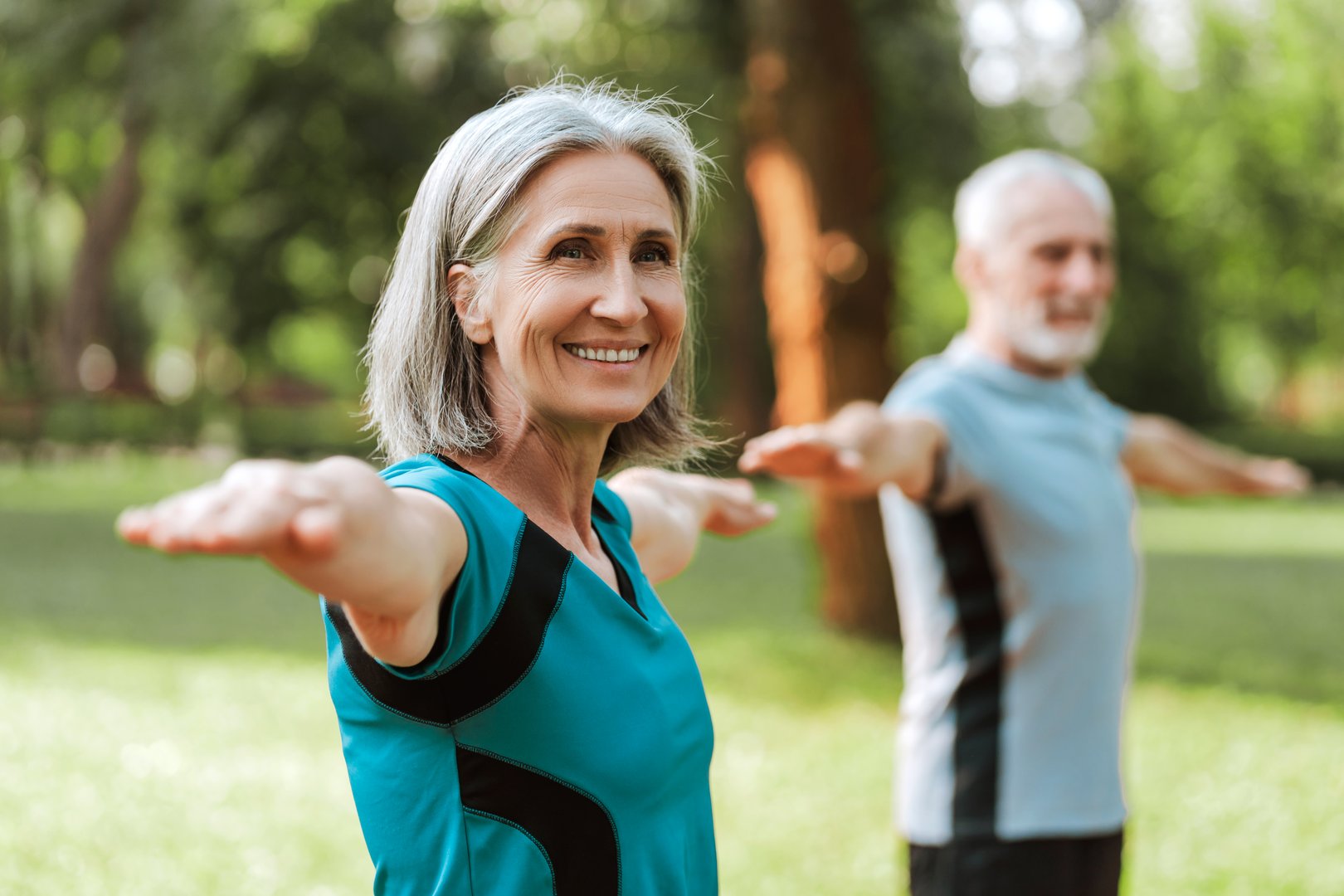 Happy senior woman stretches gracefully in a vibrant outdoor yoga class, surrounded by lush green trees and warm sunlight. Healthy lifestyle concept