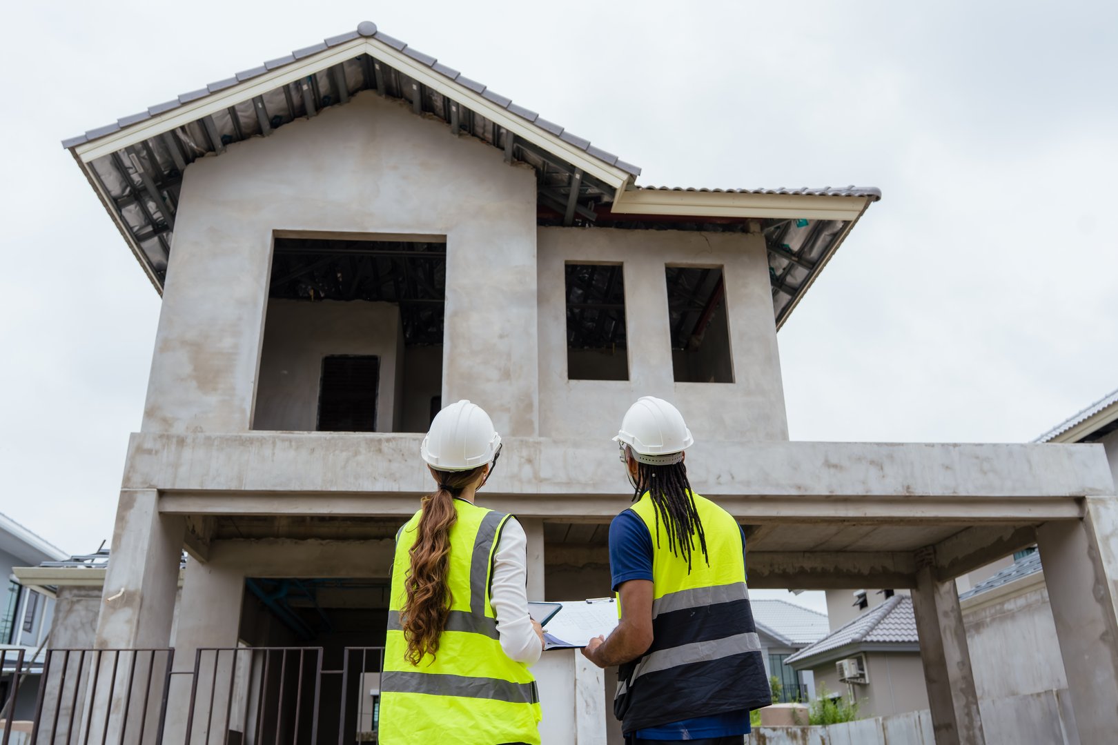 Two construction workers wearing safety gear and holding clipboards. One of them is wearing a yellow vest