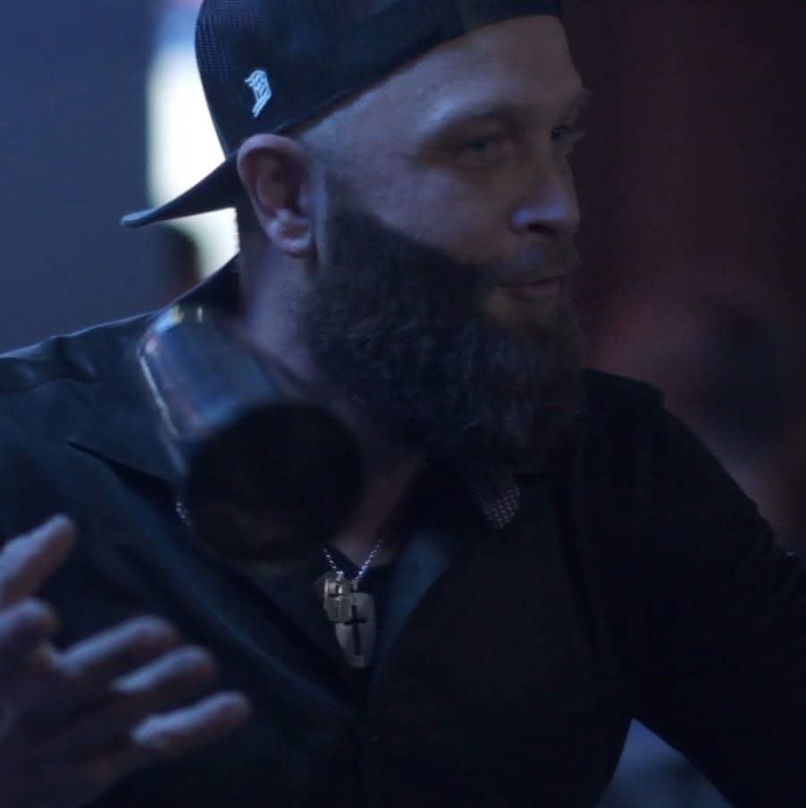 Man with a beard wearing a cap and a cross necklace, performing a flair bartending trick with a shaker.