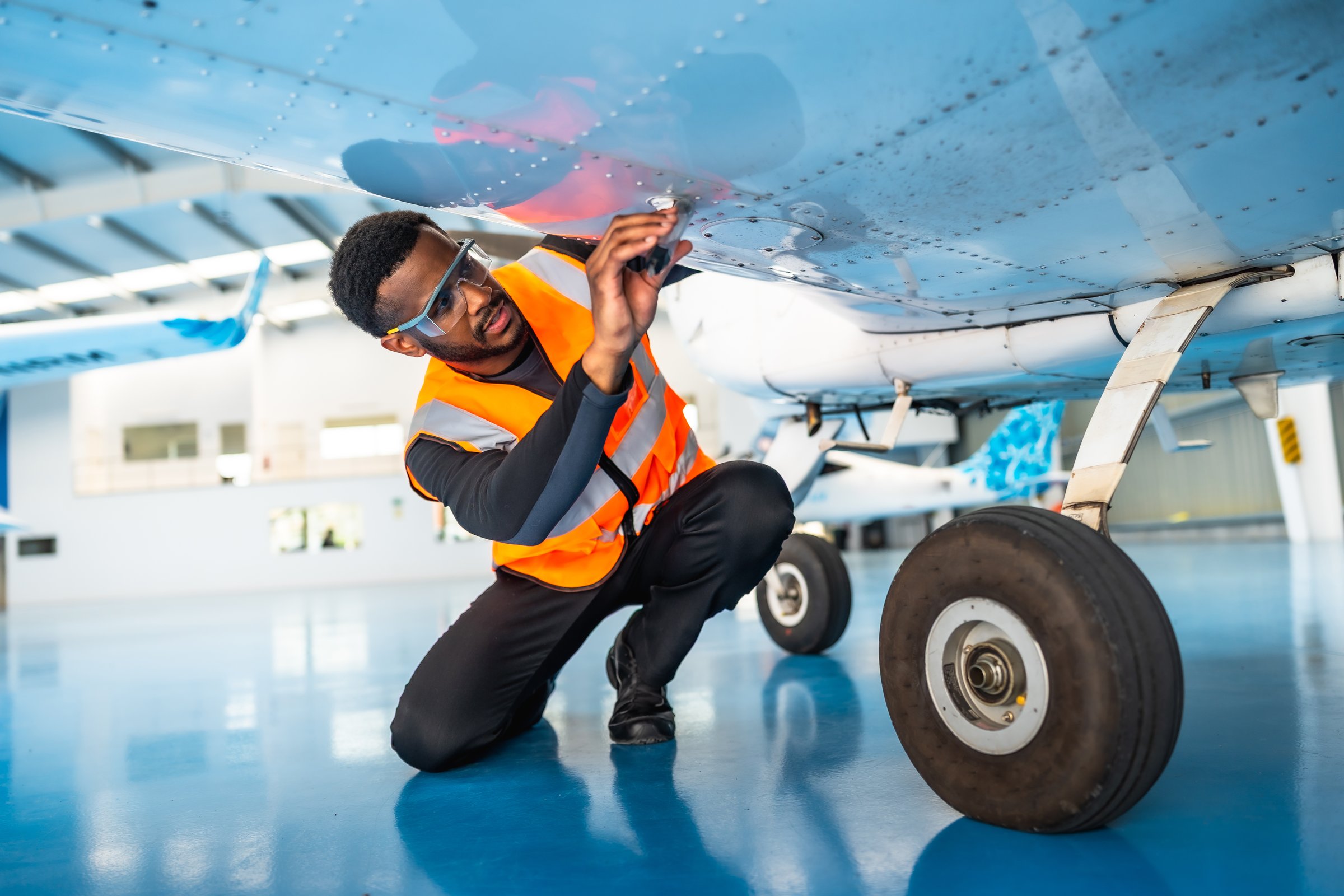 Aircraft maintenance engineer wearing safety glasses and a high-visibility vest inspects the wing of a small airplane inside a hangar