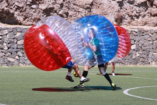 Two people on a soccer field wearing inflatable bubble suits, colliding with each other while playing bubble soccer.