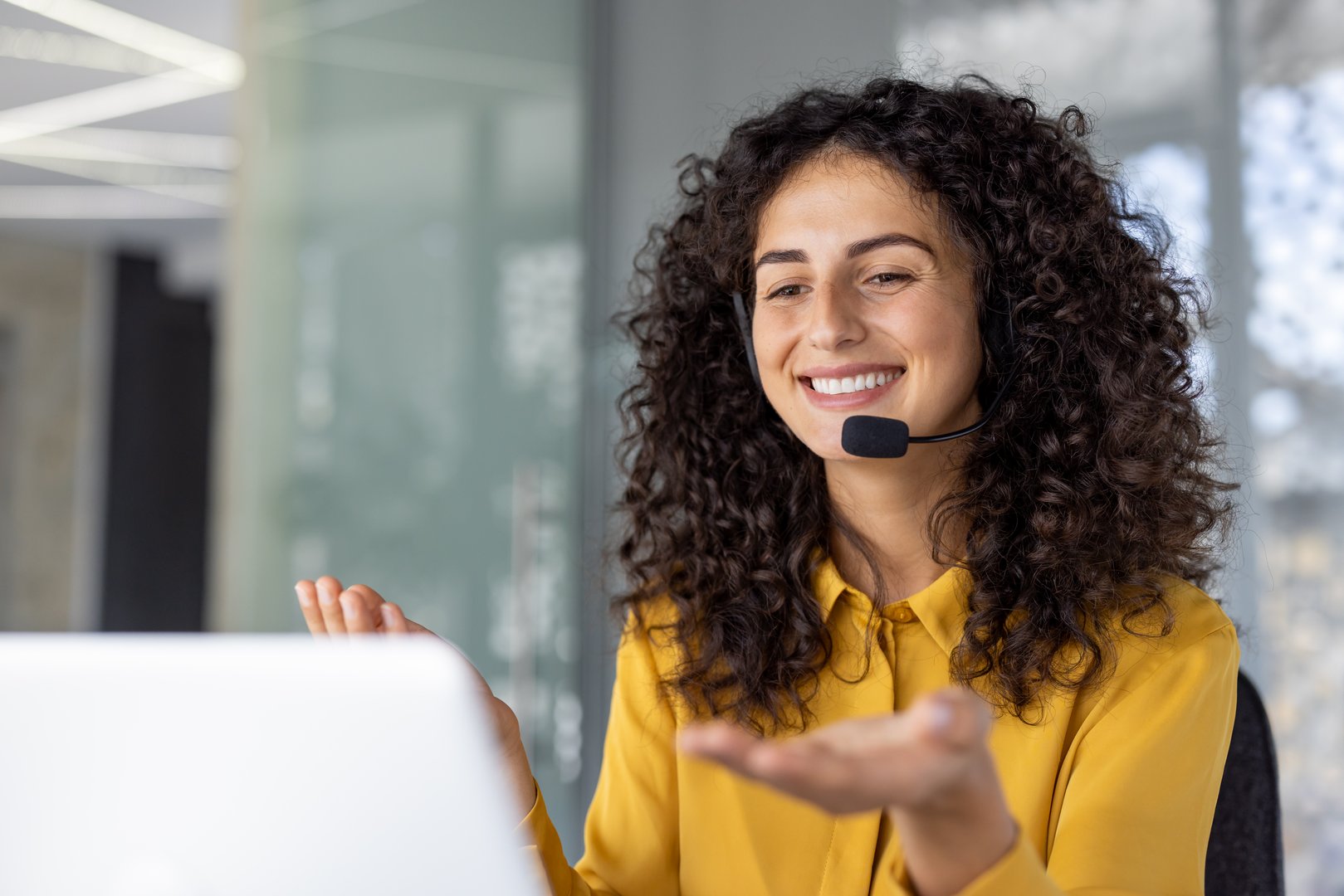 Hispanic businesswoman using headset, engaging in virtual meeting. She is in office environment, smiling and gesturing interacting. Represents communication, customer service, remote work