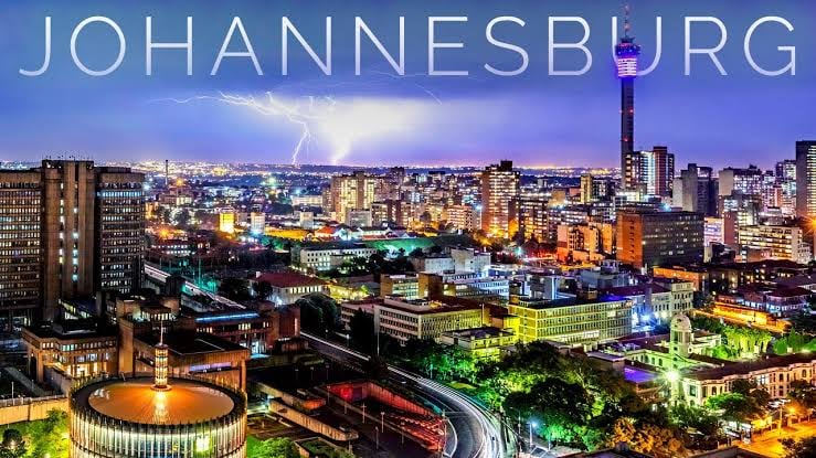 An illuminated nighttime skyline of Johannesburg with city lights, buildings, a tower, and lightning in the sky.