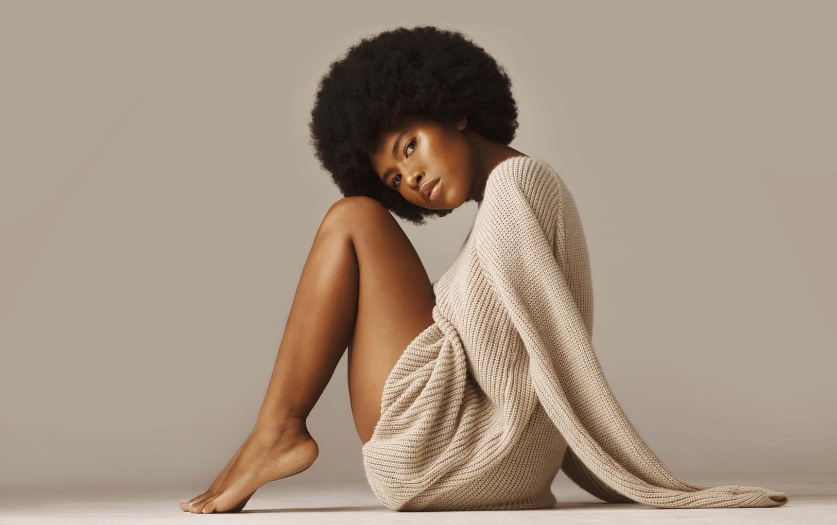Beautiful african american woman with afro wearing casual winter sweater and sitting barefoot and knees up. Attractive female with glowing skin wearing warm and cozy knitwear against brown background