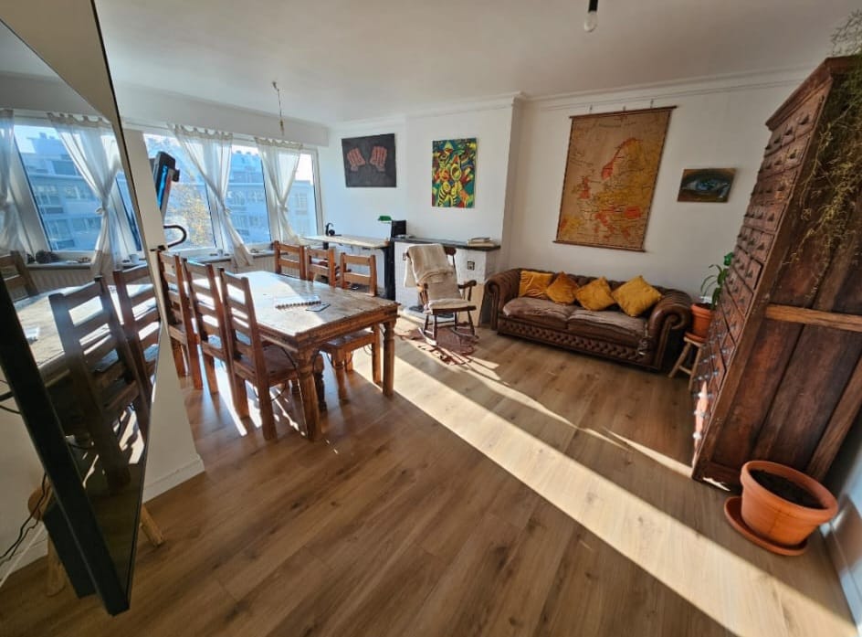 Bright living room with wooden dining table, vintage sofa, colorful wall art, and large windows with sheer curtains.