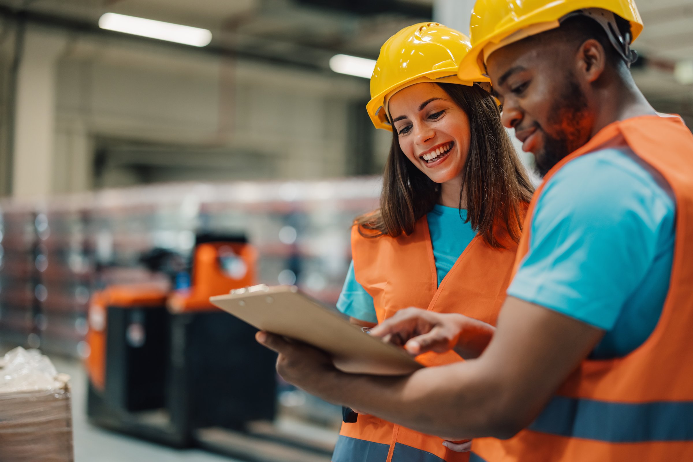 Two happy warehouse workers are smiling and looking at a clipboard while taking inventory in a large distribution center
