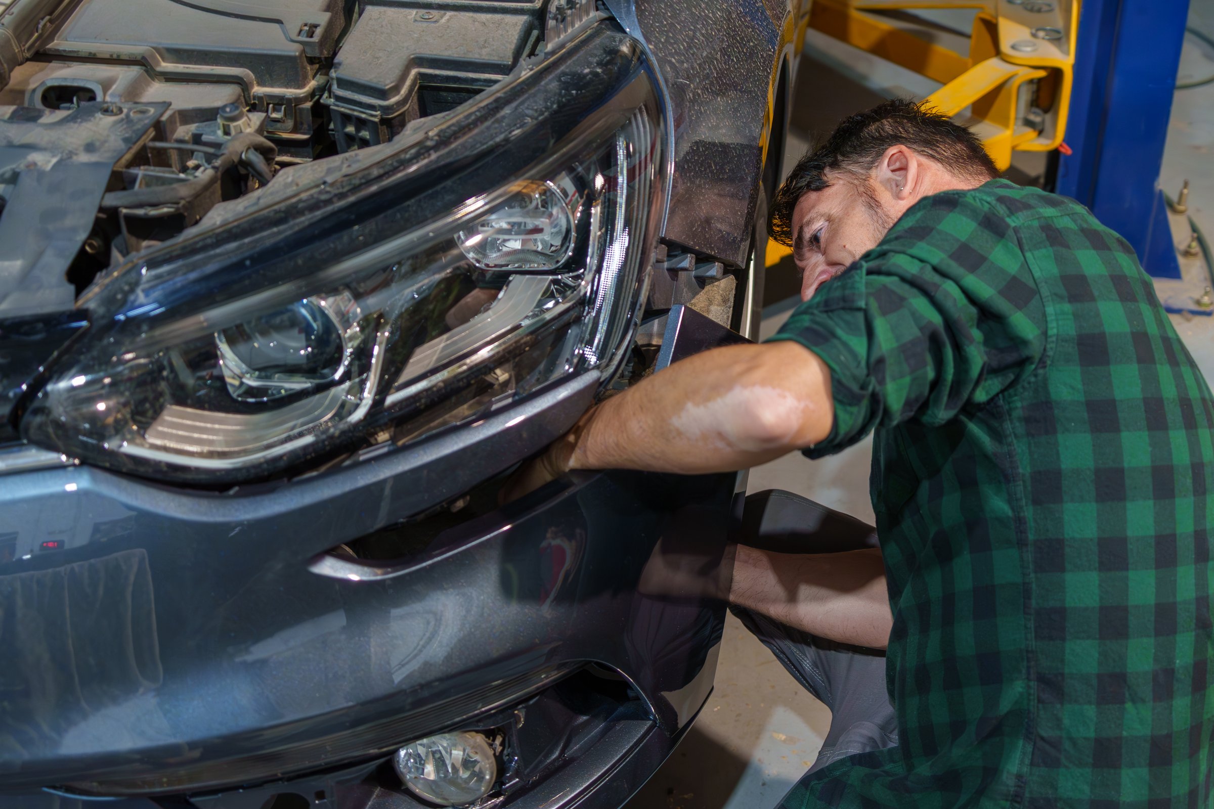 A mechanic in a green plaid shirt carefully aligns the front bumper of a car, focusing on precision and repair in a professional automotive workshop.