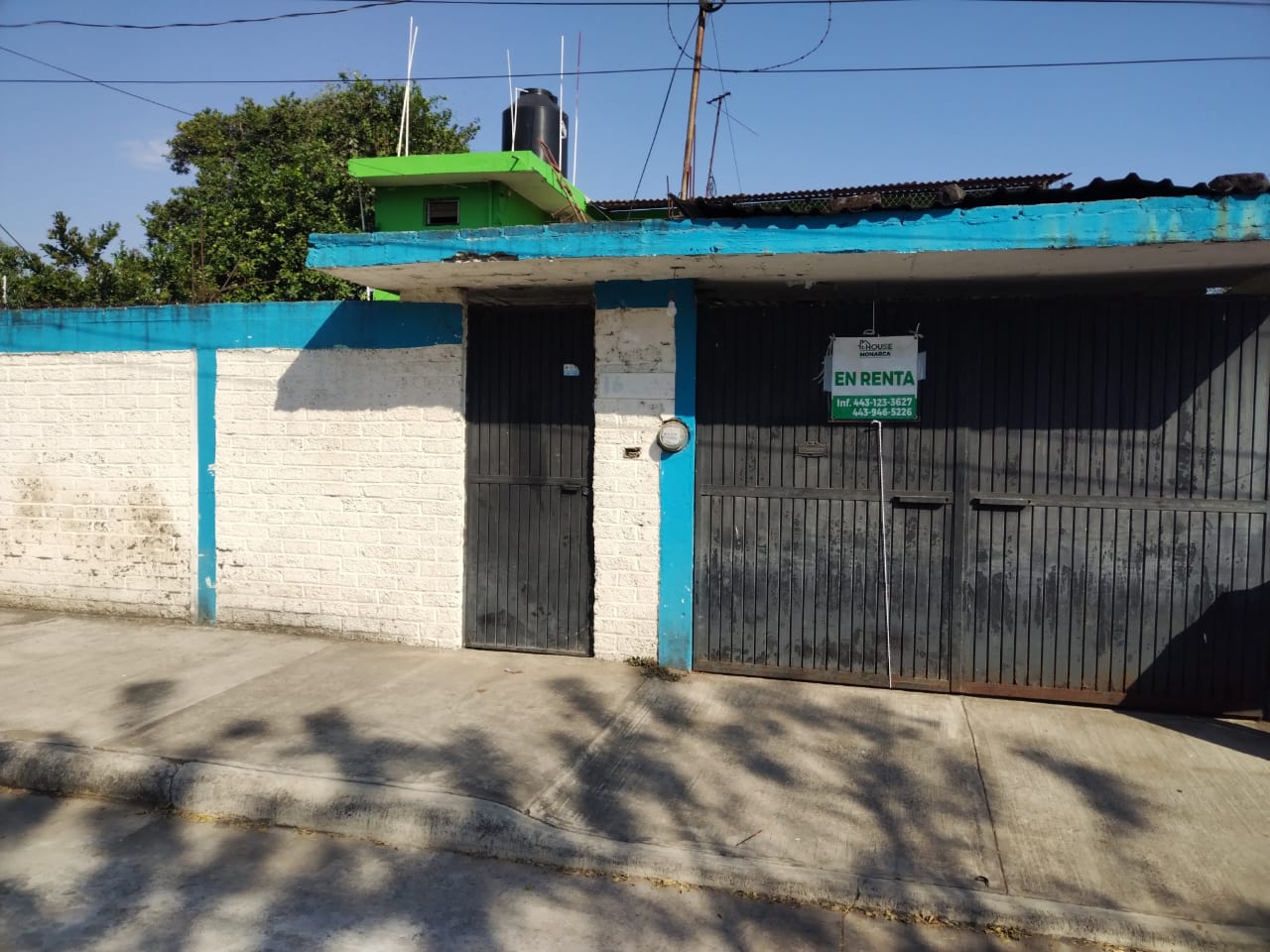 A house with a green and blue facade, featuring a "For Rent" sign on the metal gate, surrounded by trees and a clear sky.