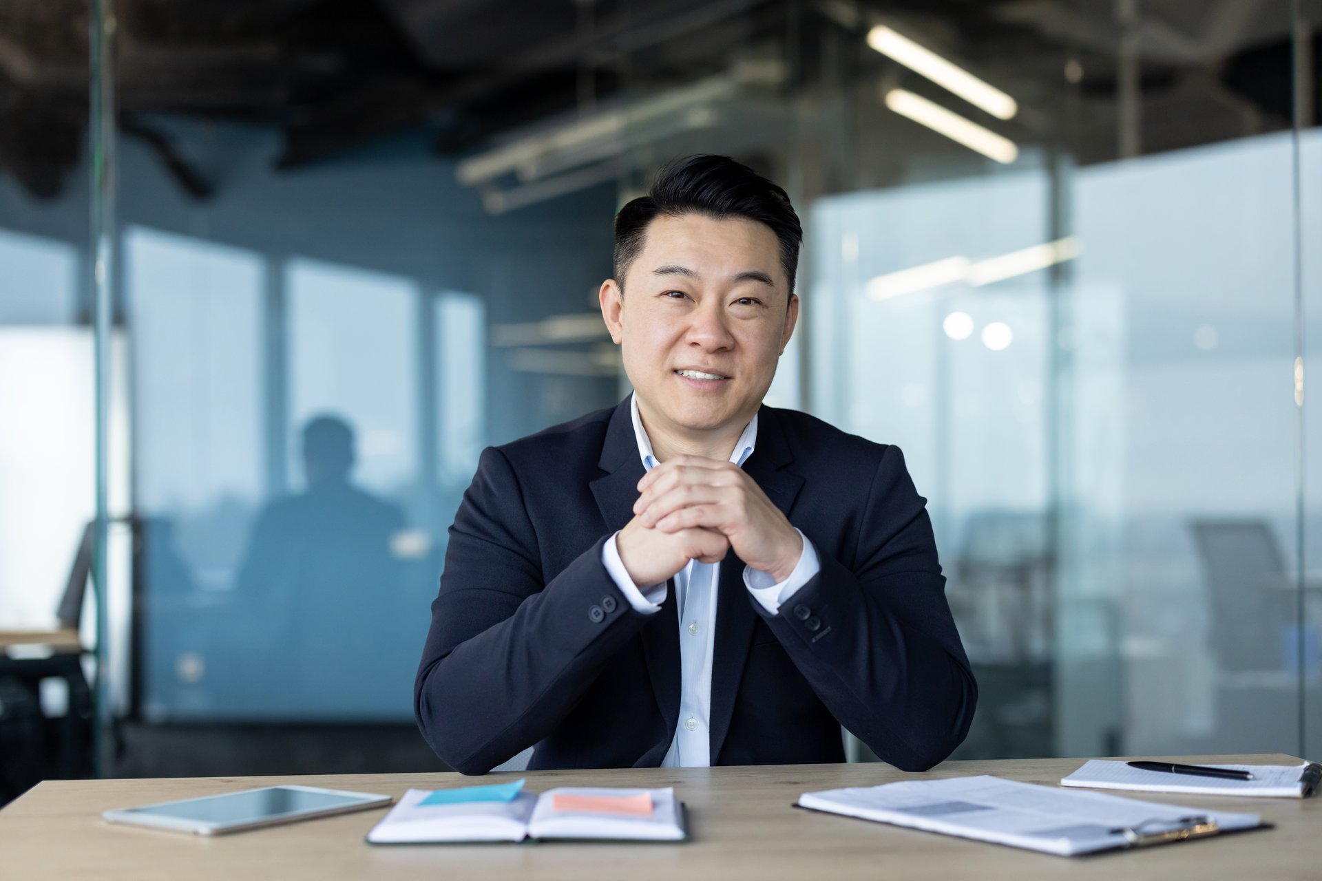Portrait of a smiling and successful young Asian businessman and director sitting in a suit at his desk and looking confidently at the camera.