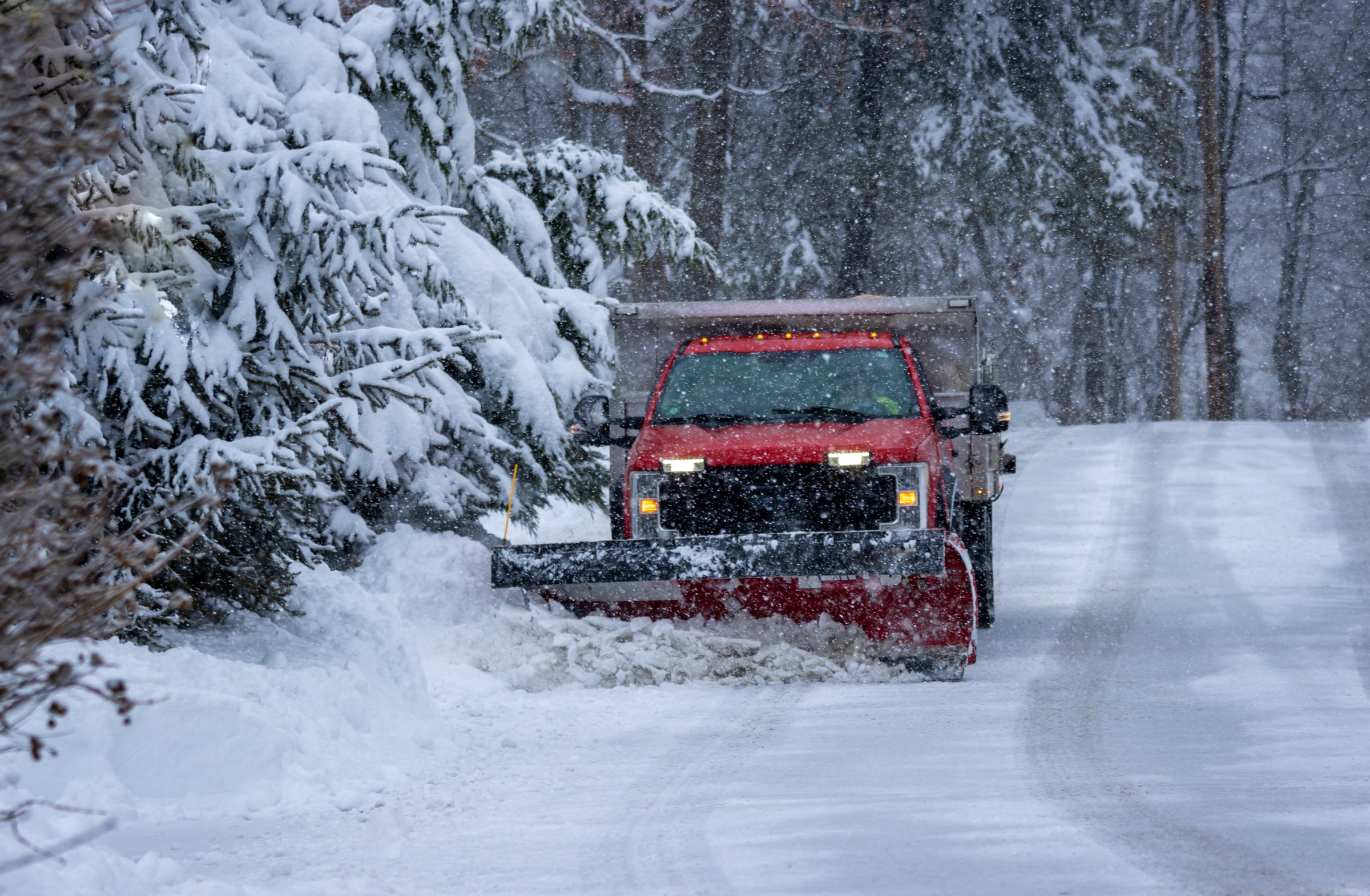 Red snow plow truck clearing snow from a residential driveway surrounded by snow-covered trees, illustrating winter snow removal services in Anchorage, Alaska.