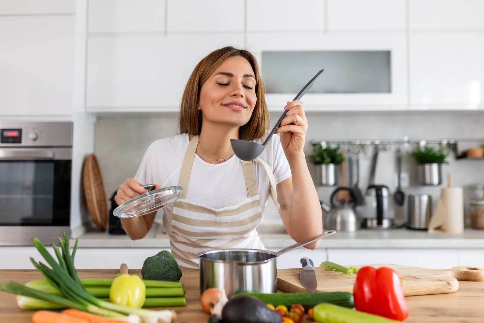 Happy Young Woman Cooking Tasting Dinner In A Pot Standing In Modern Kitchen At Home. Housewife Preparing Healthy Food Smiling . Household And Nutrition. Dieting Recipes Concept