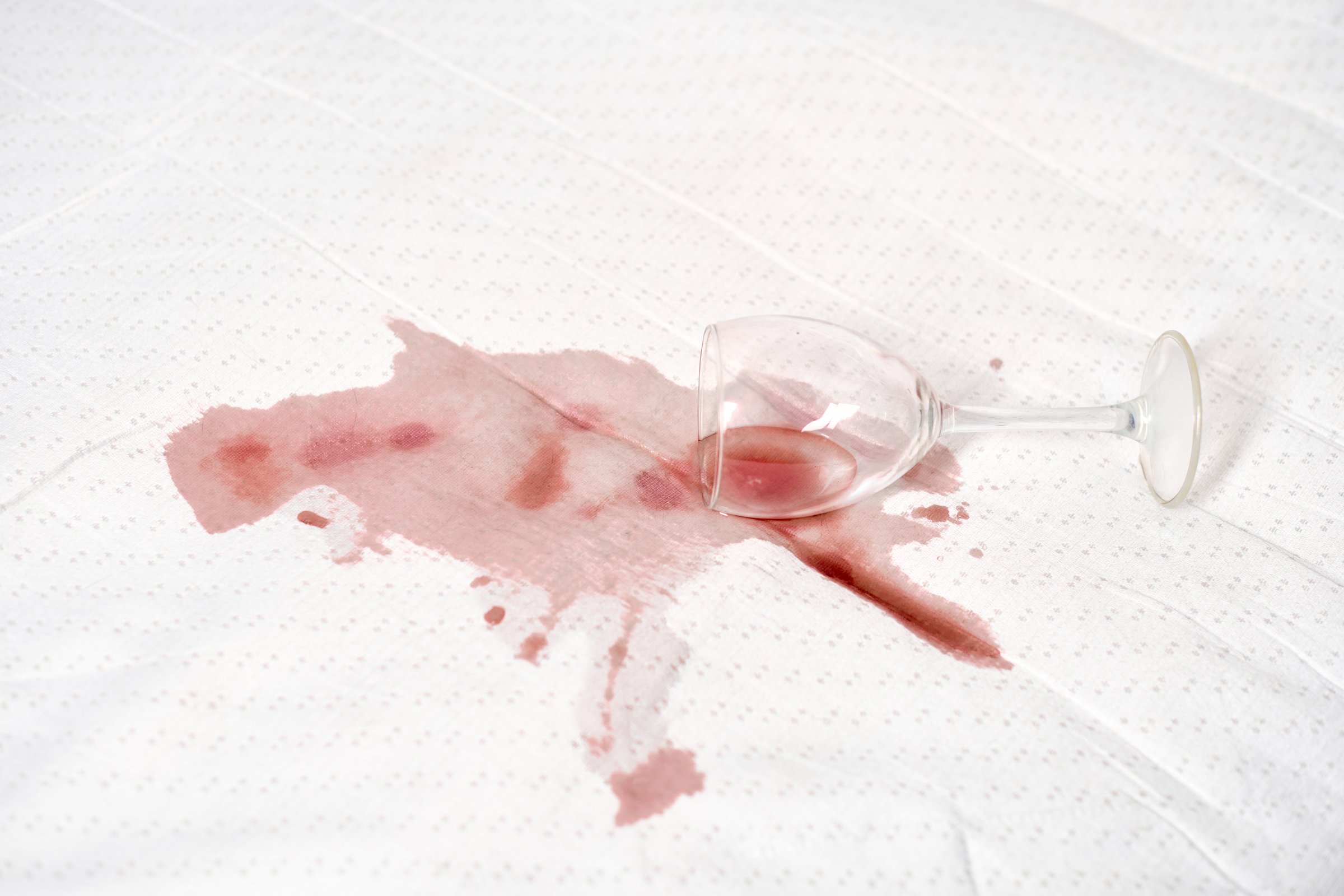 Upside-down wine glass lies on white fabric, causing red stain to spread. Natural light highlights contrast between deep red liquid and pristine fabric. Image captures moment of accidental spill