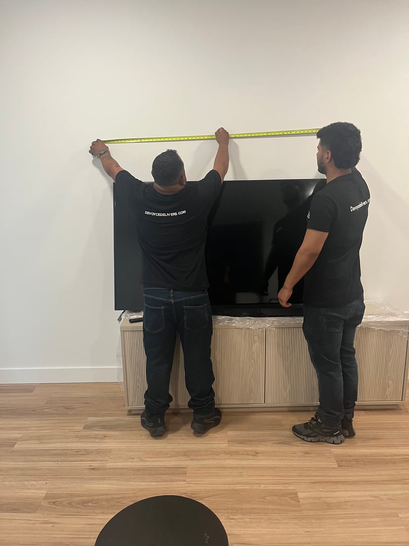 Two people measuring a wall above a mounted TV with a tape measure in a room with wooden flooring.