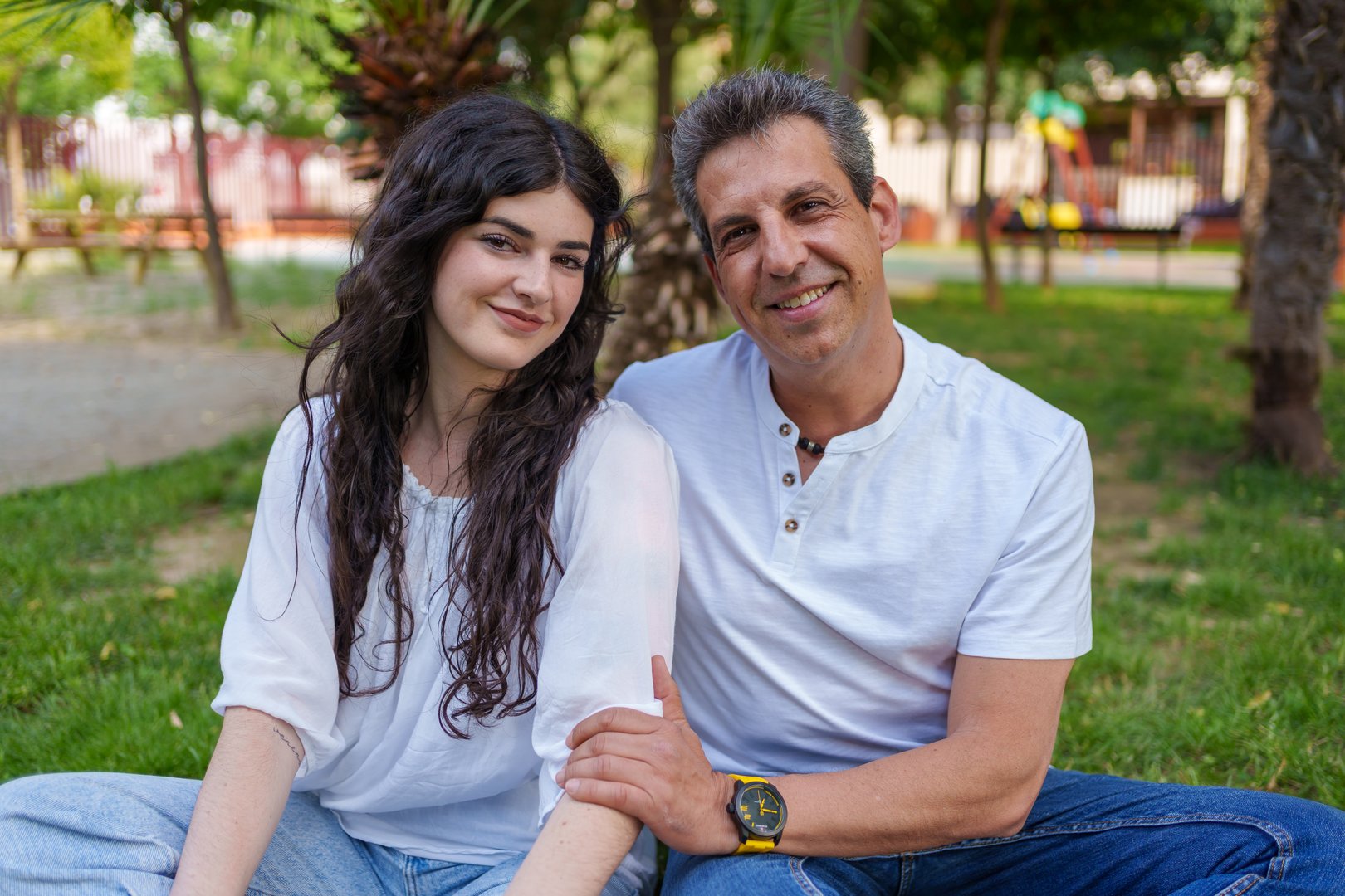 Father and daughter sitting on the grass in a park, smiling joyfully at the camera while enjoying a sunny day together, surrounded by the beauty of nature and greenery