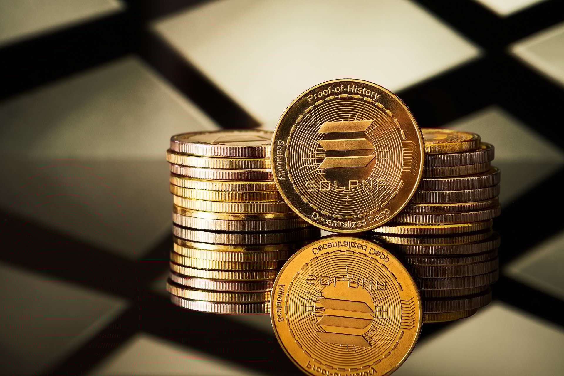 Several golden solana cryptocurrencies are arranged in a pile, reflecting off a surface with a geometric pattern in the background.