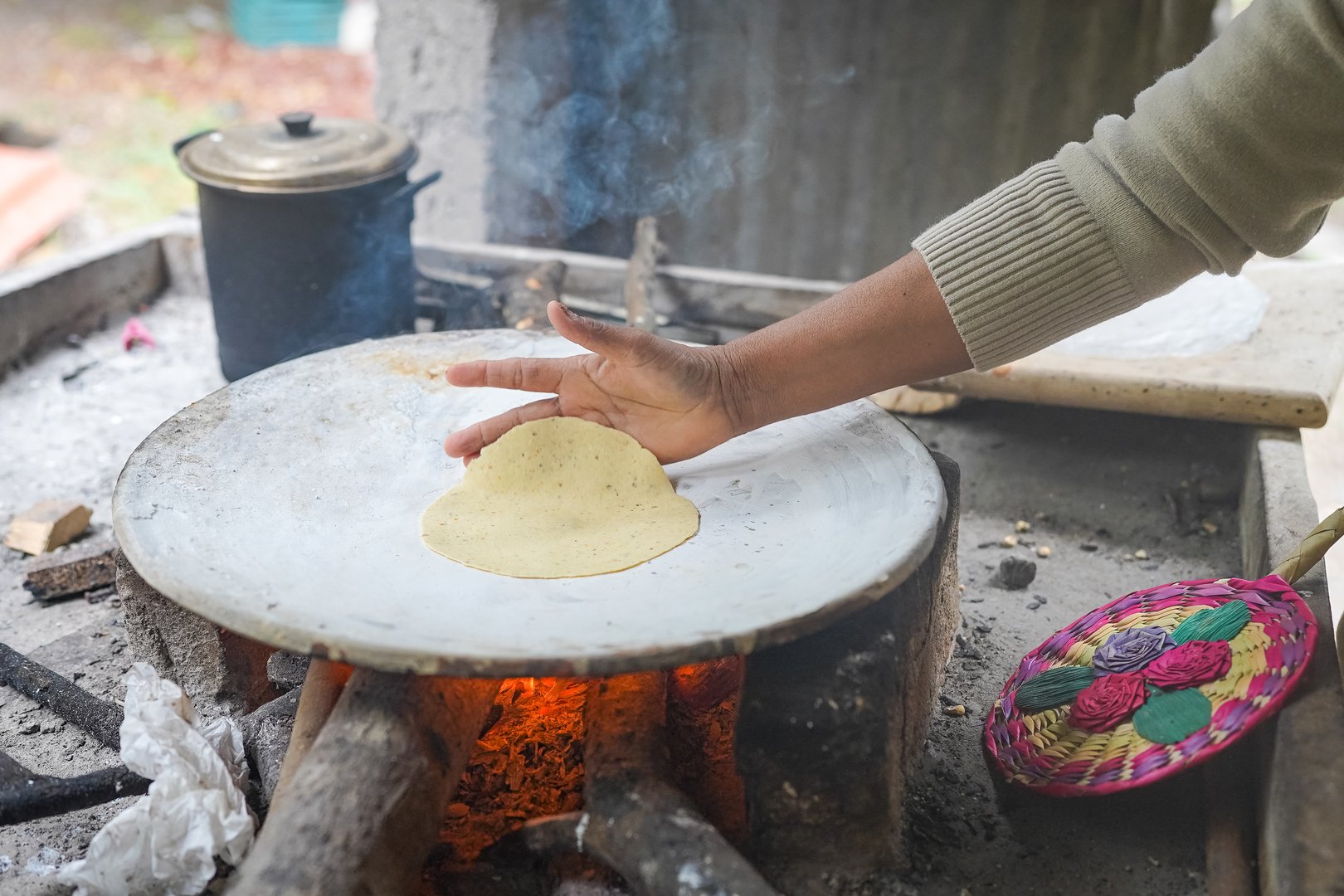 Woman's hand placing a corn tortilla on a hot comal over a wood fire in a sustainable ranch setting