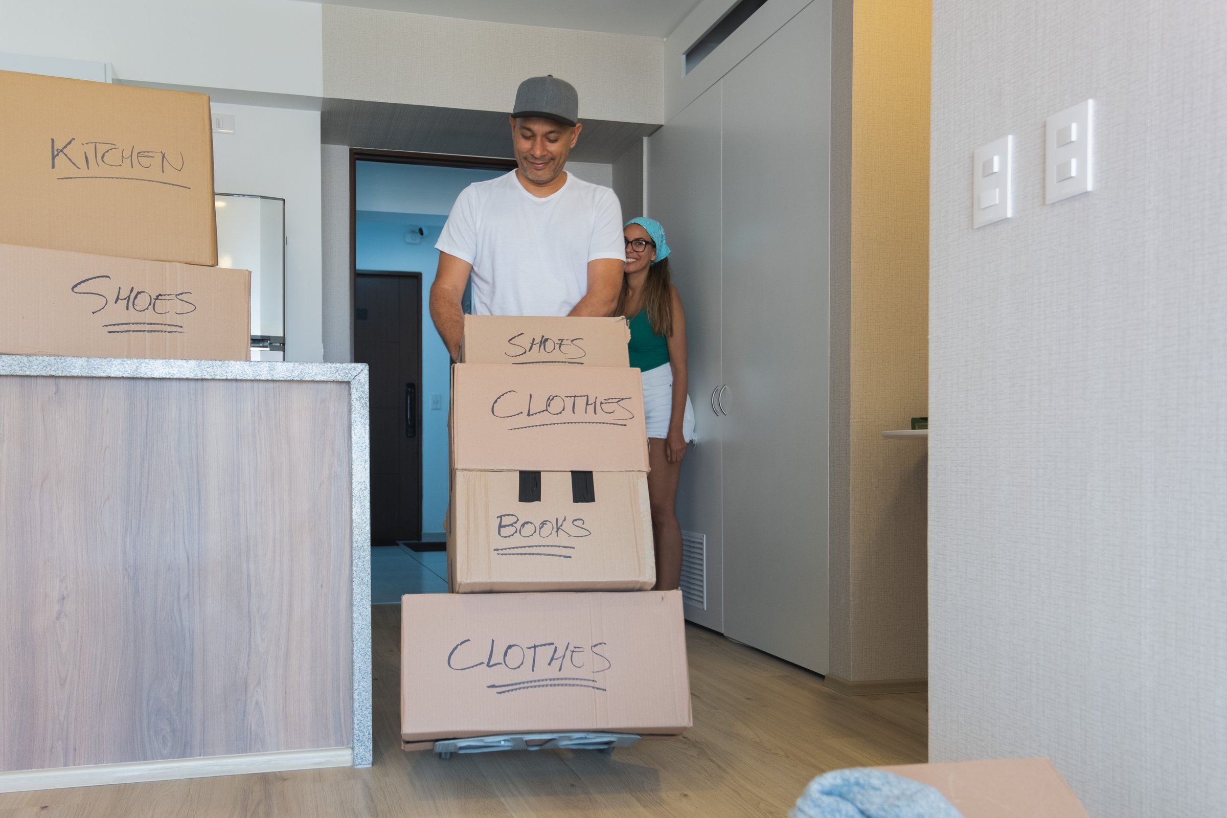 A man and a woman are moving boxes into a new apartment. The boxes are labeled with words like clothes and shoes. The man is pushing a cart with the boxes on it, while the woman is standing nearby