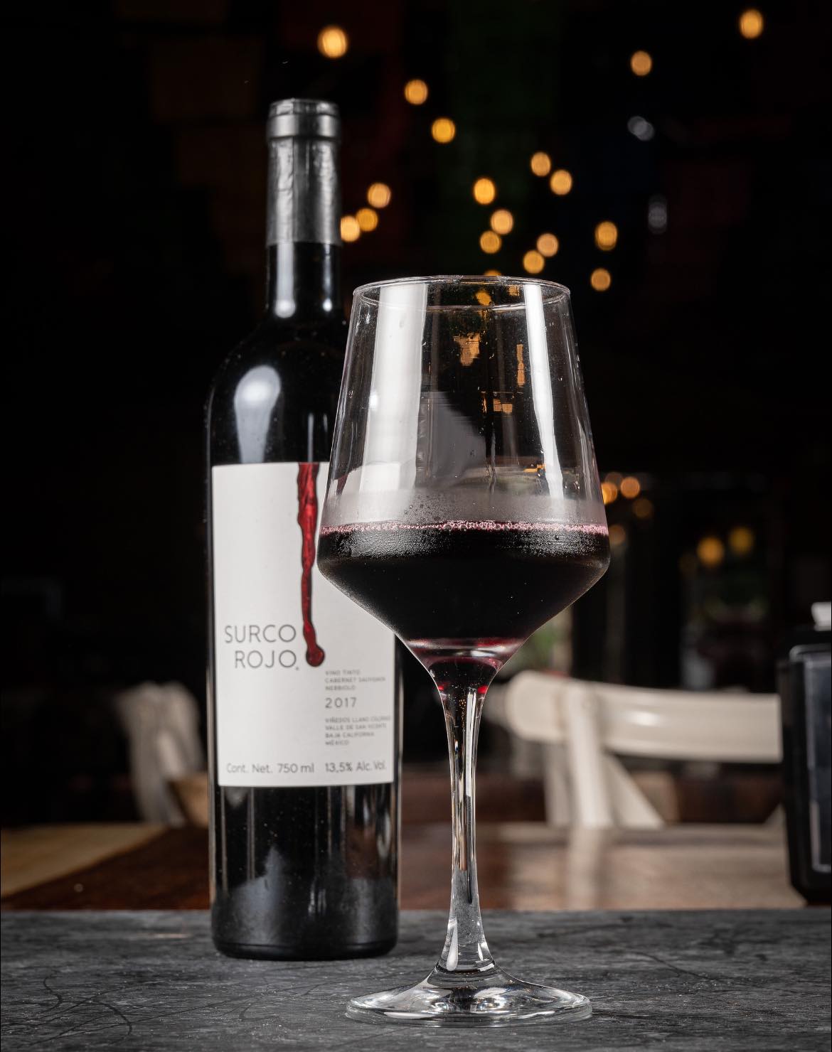 A glass of red wine in front of a bottle labeled 'Surco Rojo 2017' on a dimly lit table with bokeh lights in the background.